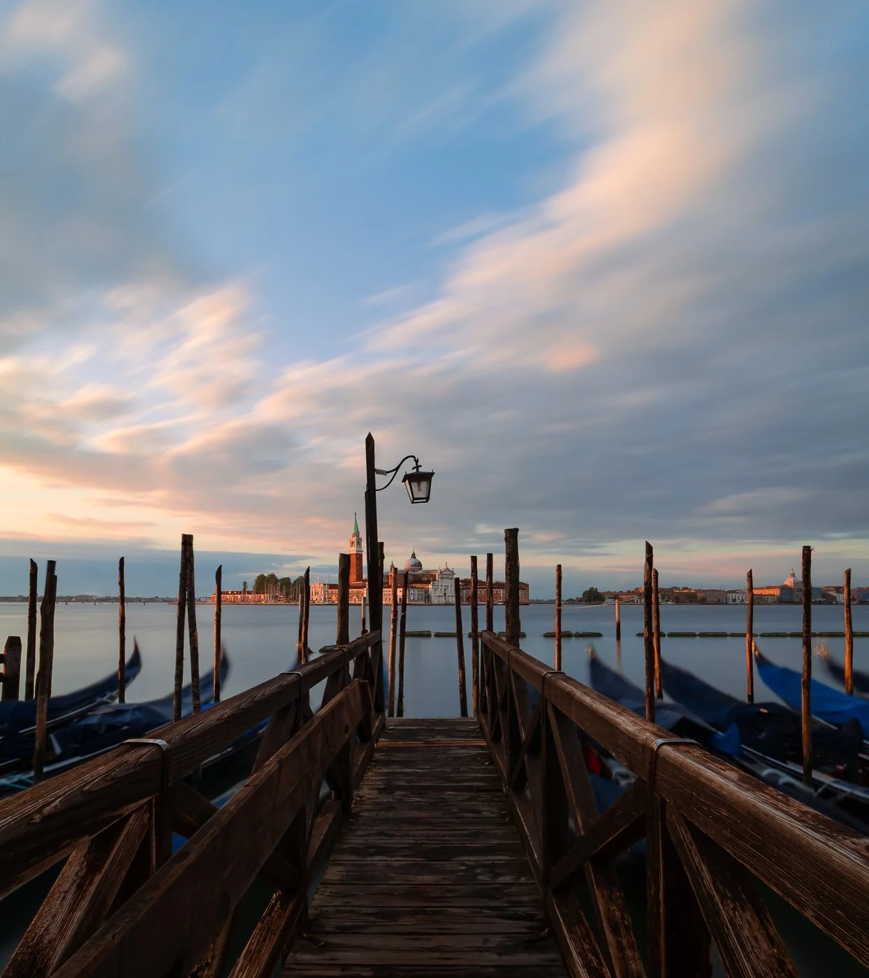 Long exposure photograph of San Giorgio Maggiore, Venice, taken at sunrise from Saint Mark’s pier. Wooden railings and blurred gondolas frame the scene in the foreground, leading the eye toward the sunlit island across the smooth water, under streake