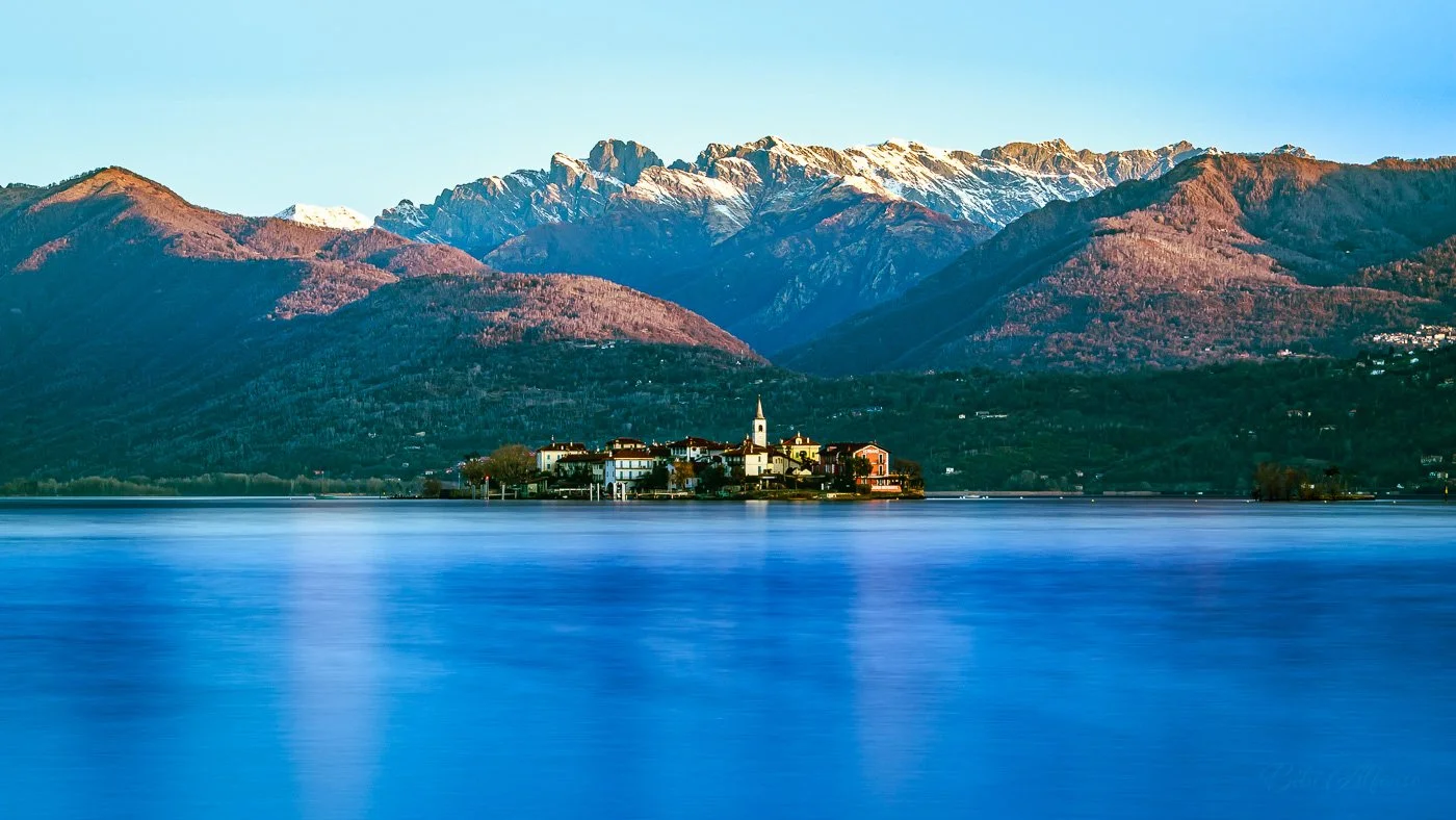 Long exposure spring shot of Isola Pescatori, Lake Maggiore, Italy, showing a lakeside town with a church steeple, calm blue water, and snow-capped mountains in the background.