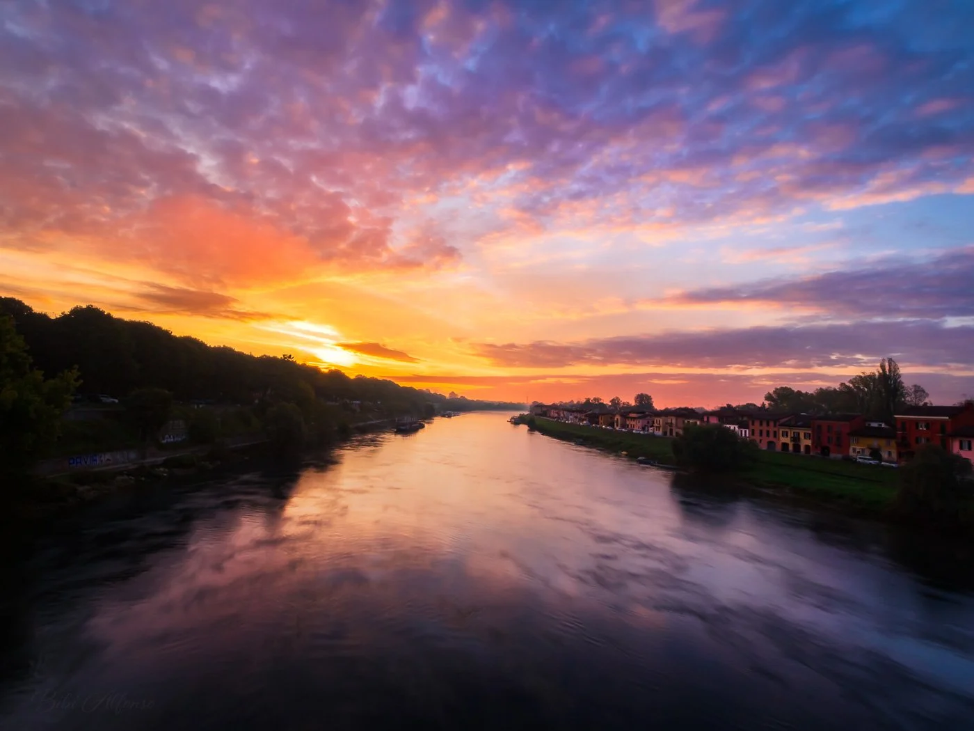Spectacular, colorful sunrise over the Ticino River in Pavia, with warm orange light illuminating the water and contrasting against the cool blue sky.