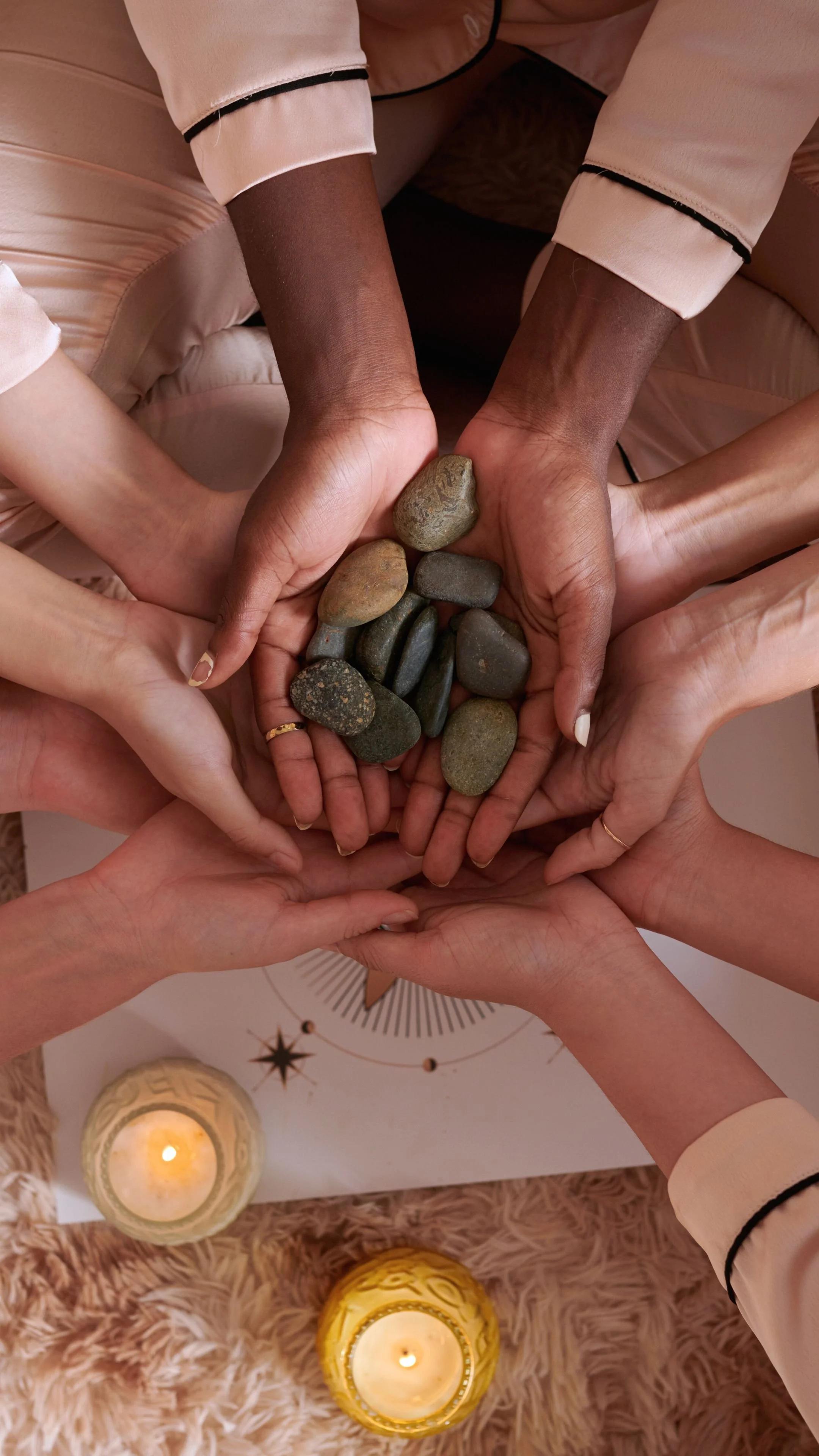 Multiple hands of different skin tones coming together in a circle, holding a collection of smooth stones, with lit candles and a white surface with black and gold designs beneath.