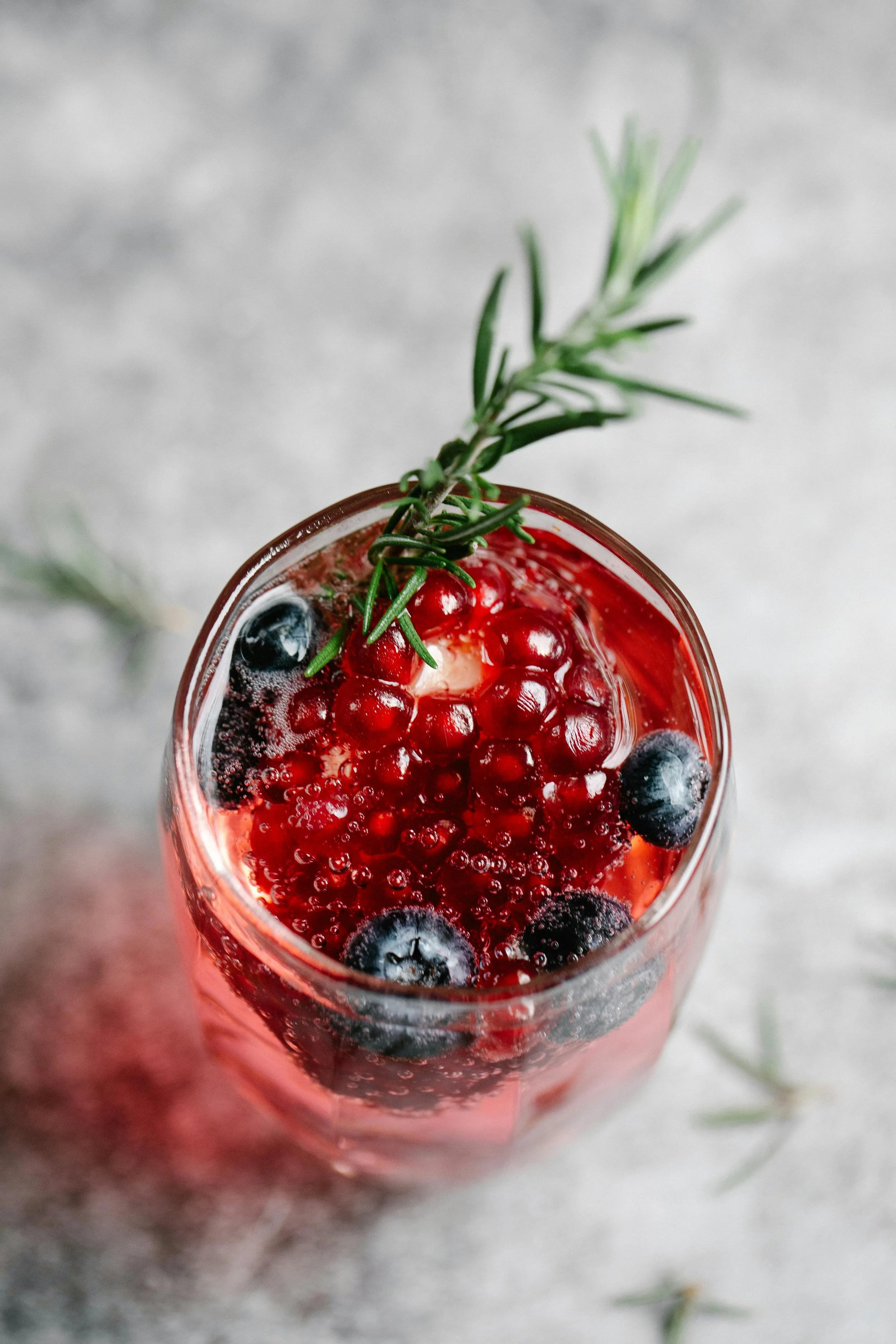 A glass of sparkling berry cocktail garnished with a sprig of rosemary, containing blueberries and red currants.