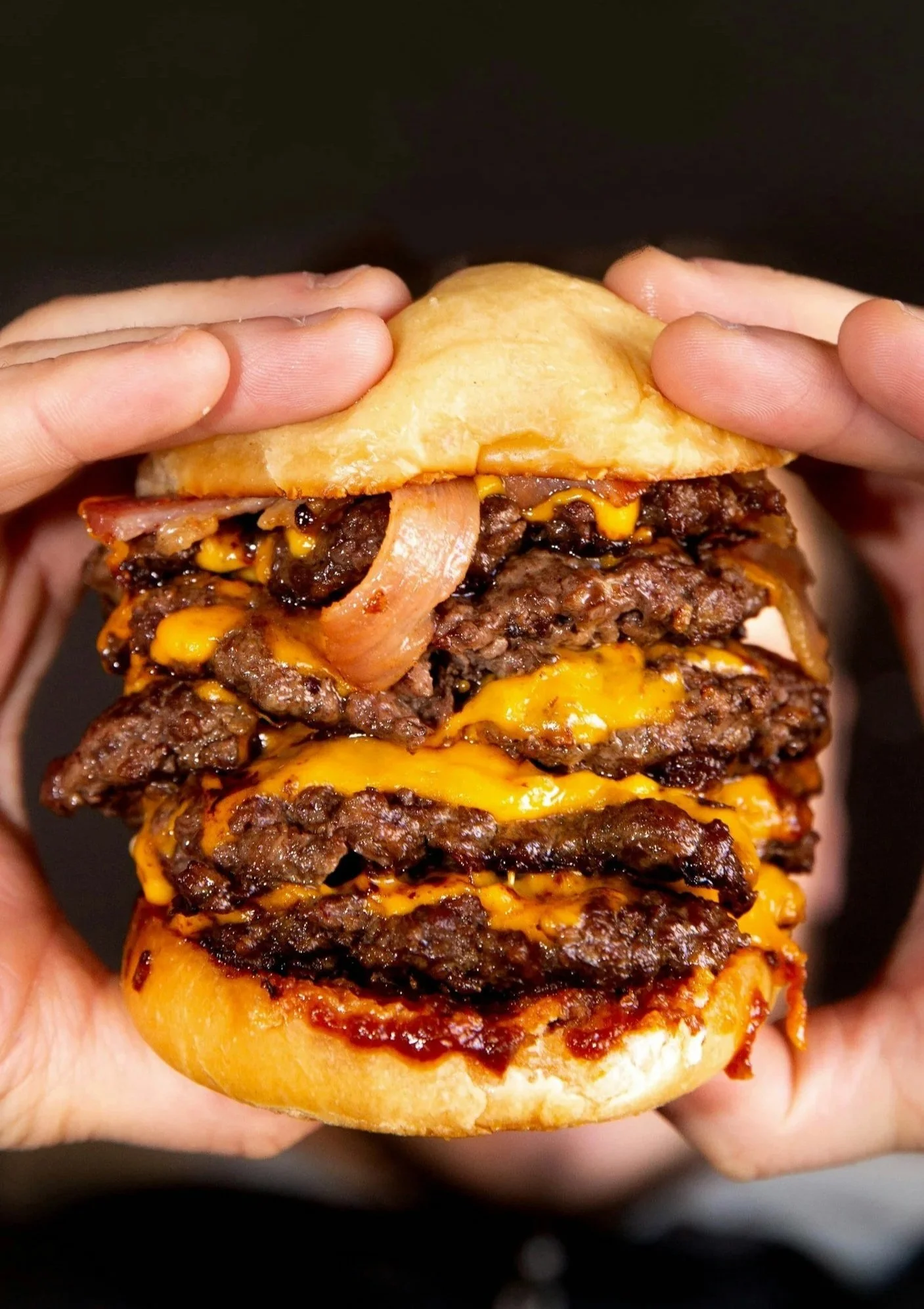 Close-up of a large smash burger with multiple beef patties, cheese, onions, and a bun, being held with both hands.