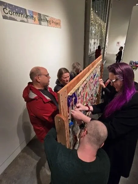 Group of people working together on a community weaving project at an indoor event, with a sign in the background that reads 'Community Weaving Project'.
