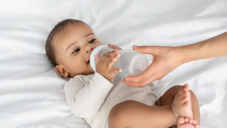 A baby lying on a bed, holding a baby bottle, being handed another bottle by an adult hand.