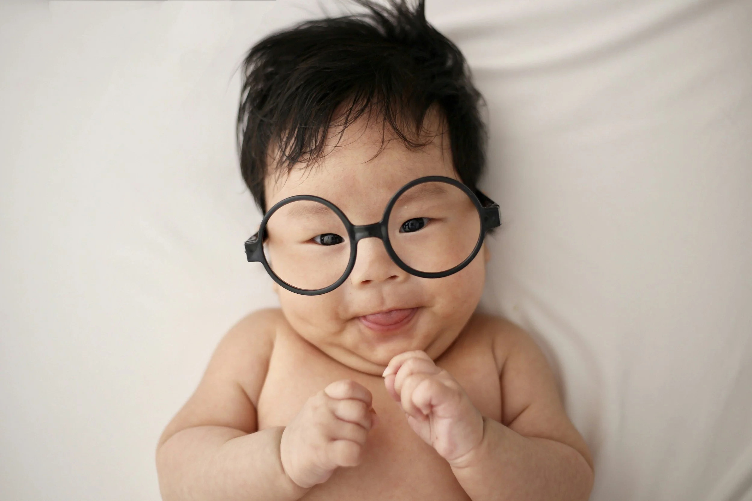 A smiling baby with glasses lying on a white surface, with hands near face and tongue slightly out.
