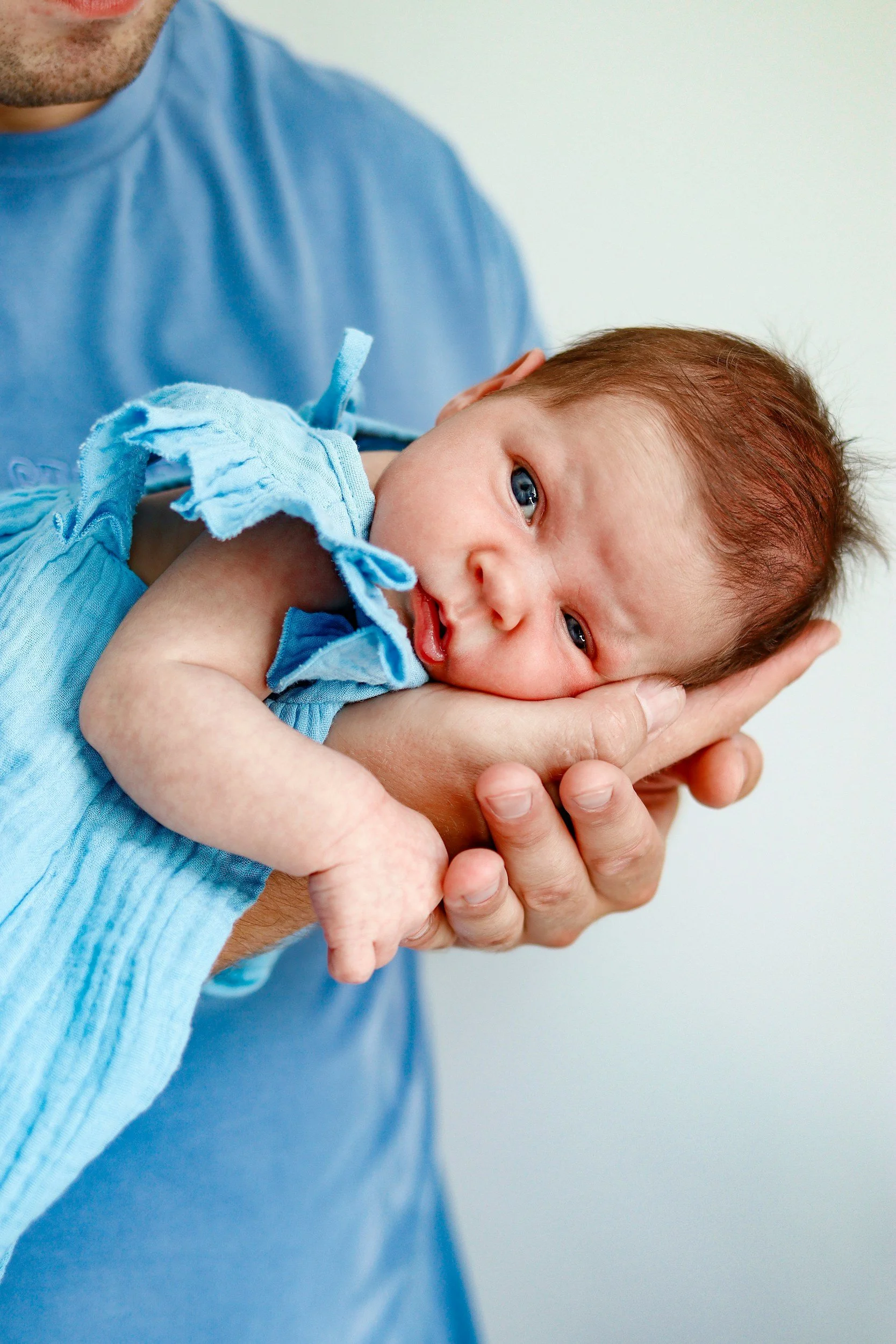 A newborn baby with reddish hair is resting on an adult's hand, looking at the camera with blue eyes. The baby is wearing a light blue outfit.