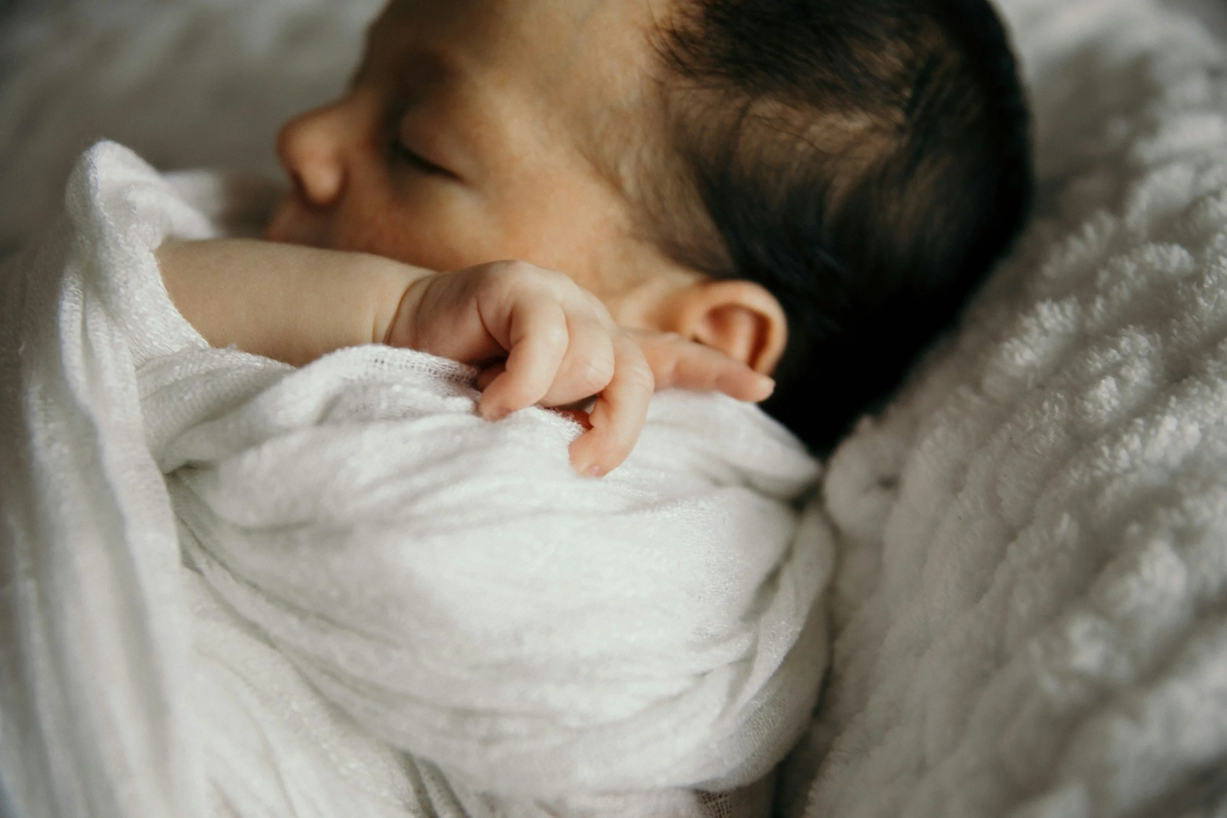 Close-up of a sleeping infant, wrapped in white cloth, lying on a soft, textured blanket.
