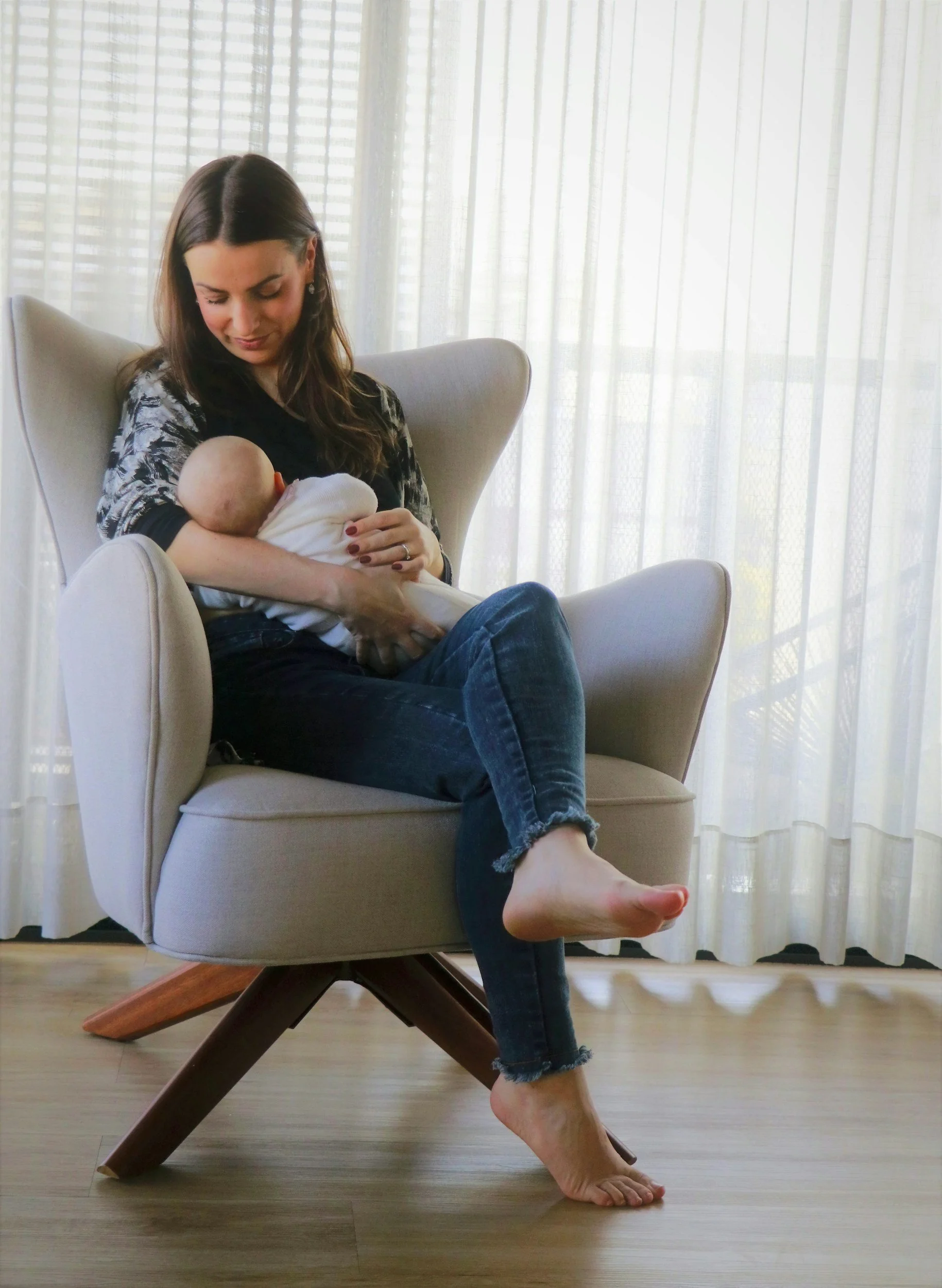 A woman sitting in a modern armchair, breastfeeding a baby, with light filtering through sheer curtains in the background.
