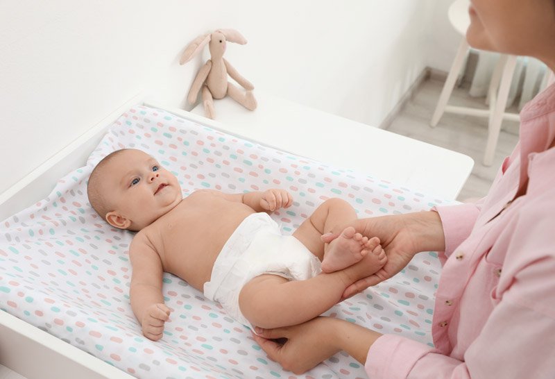 A baby lying on a changing pad on a white changing table, with a caregiver gently holding the baby's feet. A stuffed bunny toy is on the table in the background.