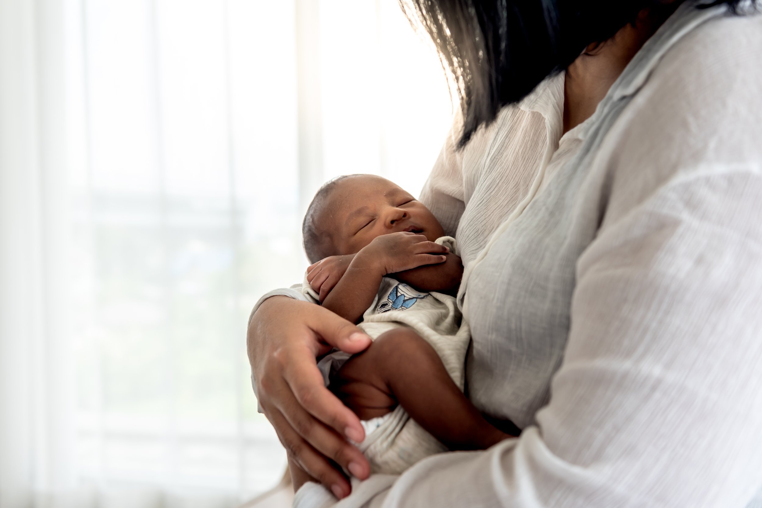 A person holding a sleeping baby close to their chest inside a well-lit room with white curtains.