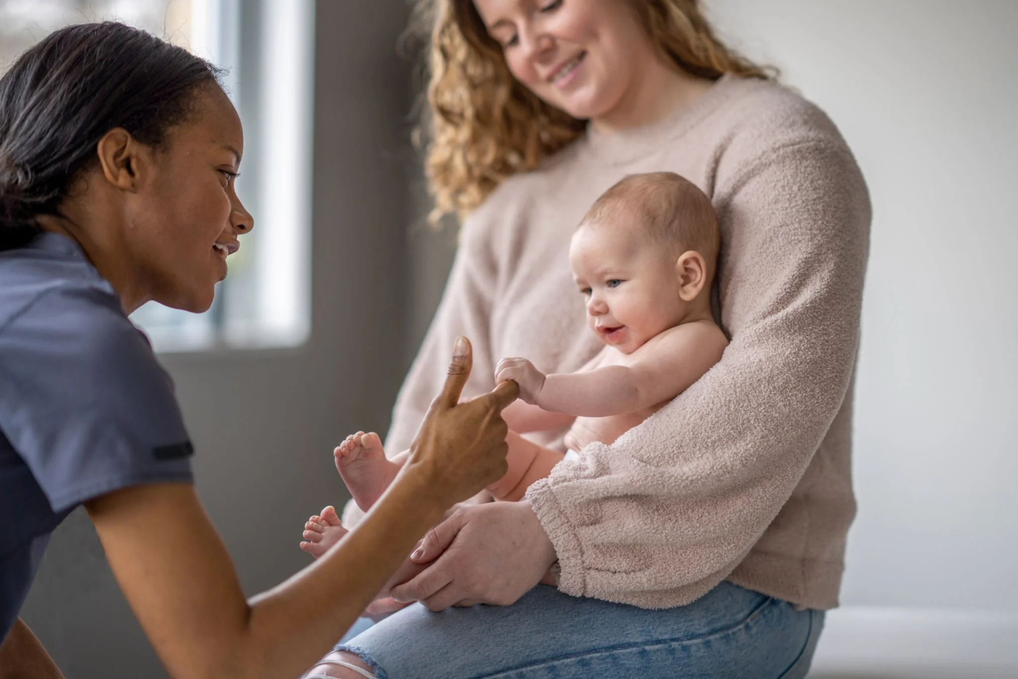 A woman with dark hair interacting with a smiling baby girl, who is seated on a woman's lap and reaching out to touch a nurse's finger. The woman with curly hair is holding the baby, and they are indoors near a window.