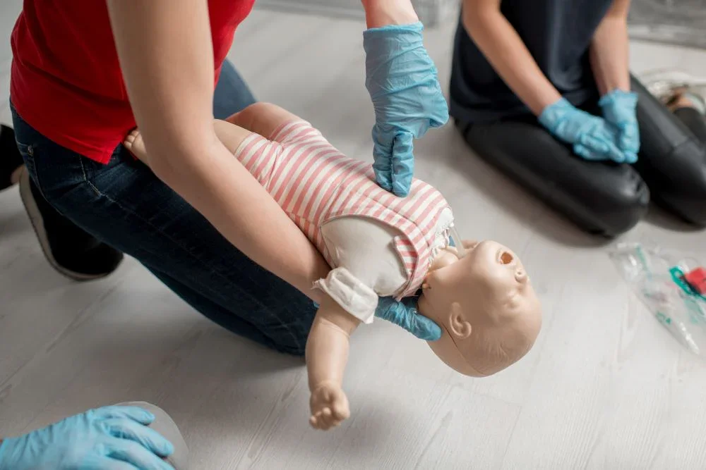 Two people practicing infant CPR on a baby mannequin, with one person kneeling and pressing on the mannequin's chest, both wearing blue gloves.