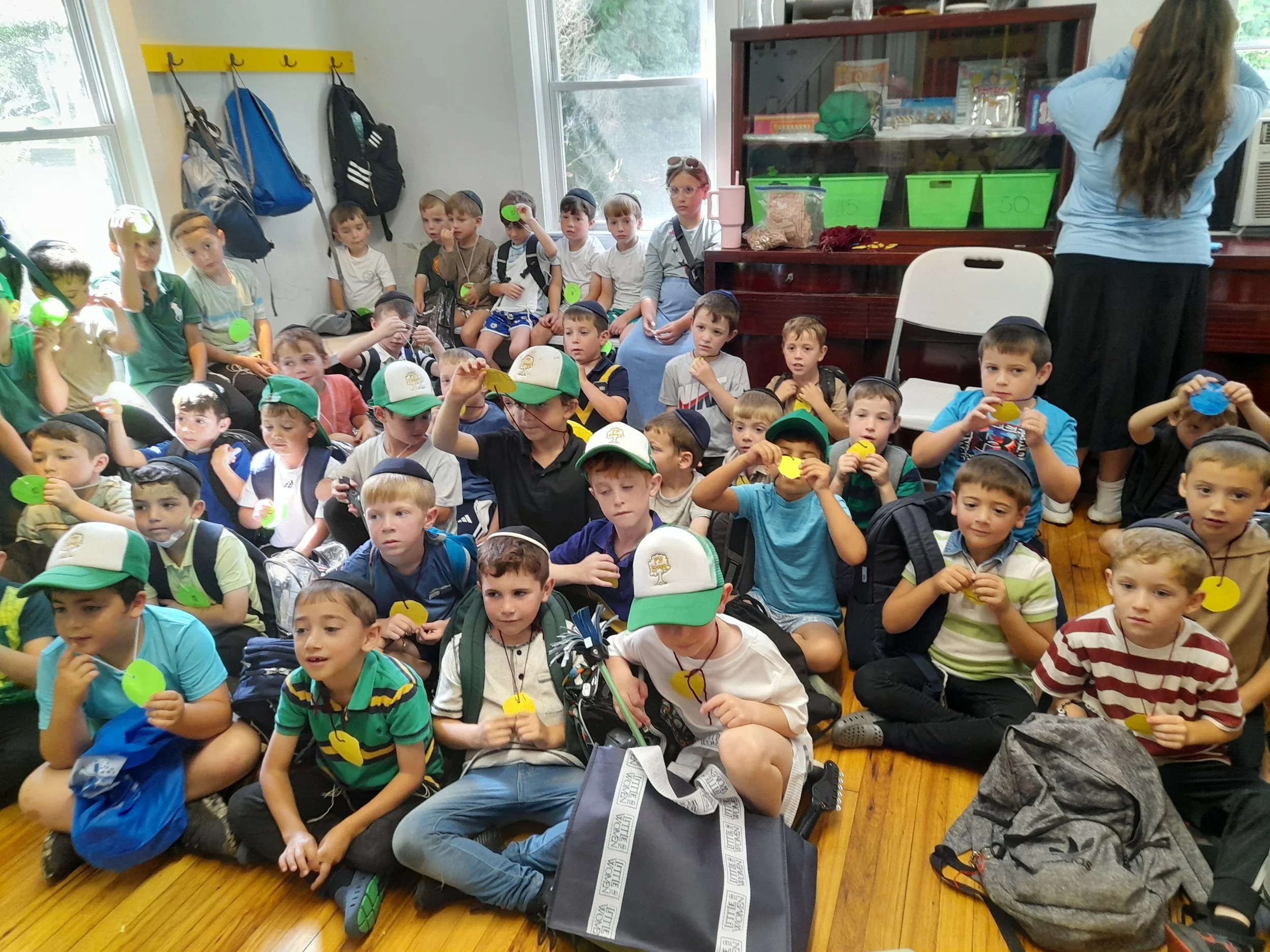 A group of young children sitting on a wooden floor, holding yellow and green name tags, with backpacks beside them. Some children are wearing hats and are seated in a classroom with a woman standing at the back.