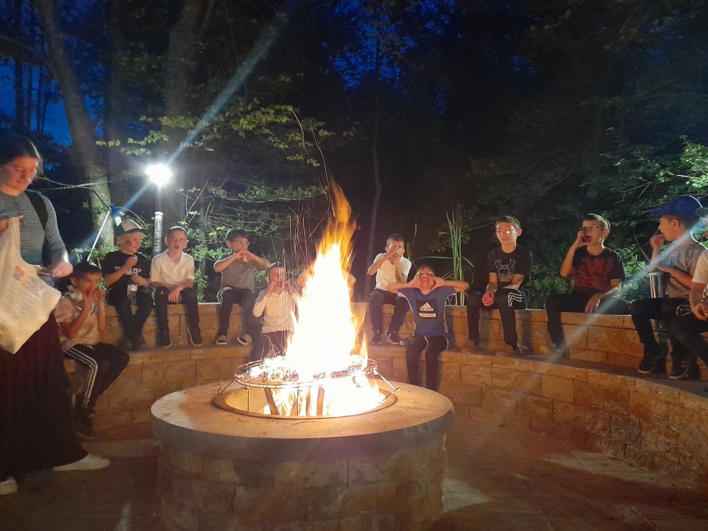 Group of kids sitting around a campfire, eating and chatting, with one adult supervising.
