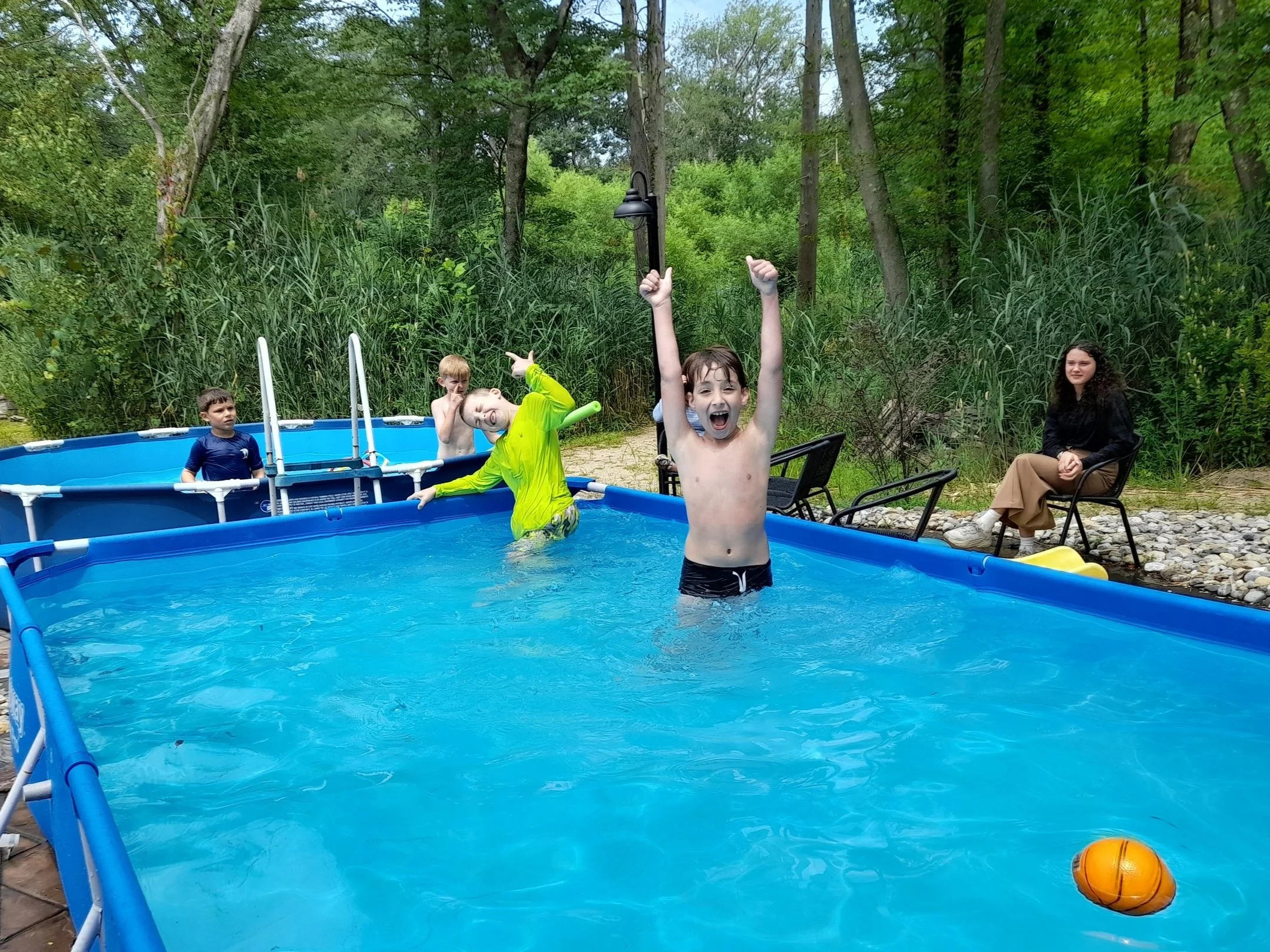 Children playing in an above-ground pool, surrounded by trees, with a person watching poolside.
