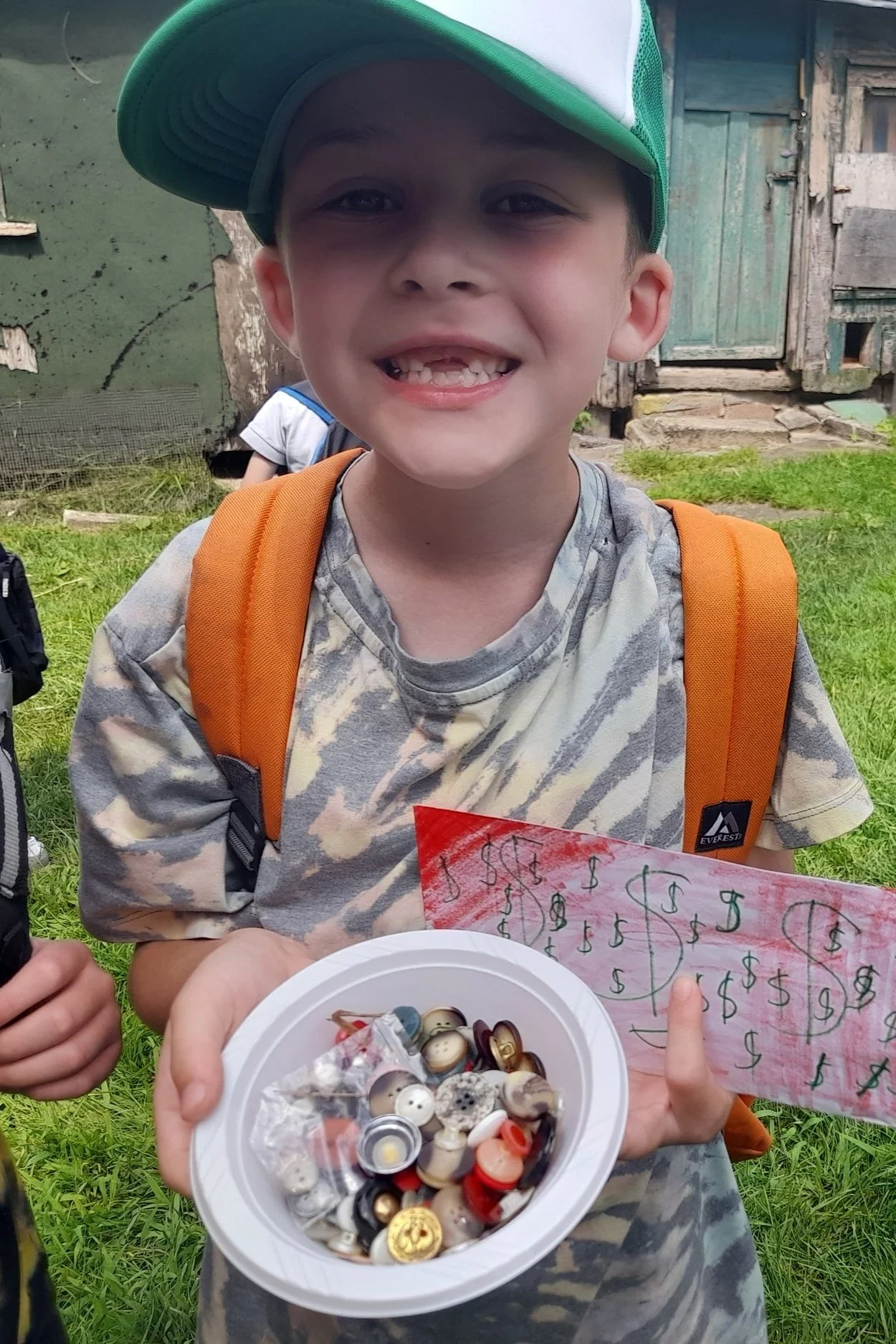 Child in camo shirt and green cap holding a bowl of buttons and handmade money.