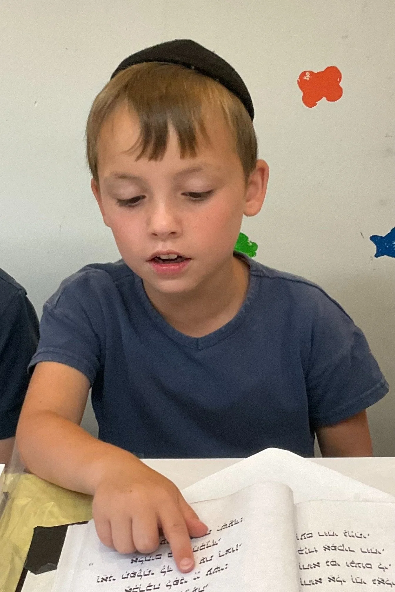 Young boy wearing blue shirt and black cap reading a book with Hebrew text.