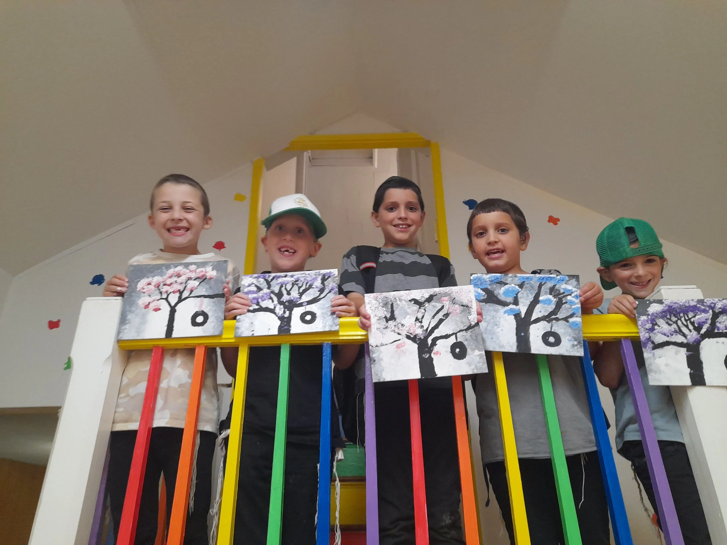 Smiling children each holding paintings of colorful trees, standing behind a rainbow-colored railing.