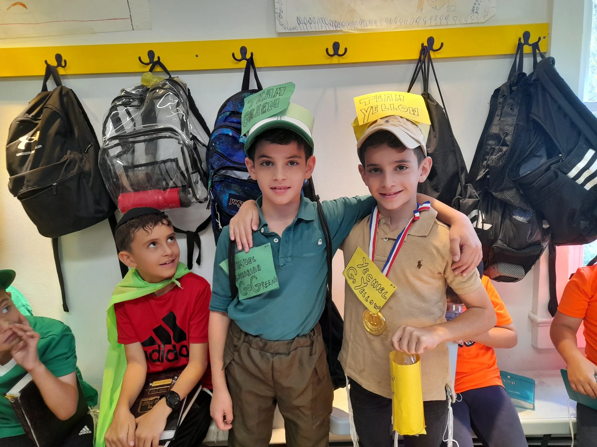 Three boys celebrating a sports achievement, with medals and hats labeled 'Team Yellow' and 'Team Green,' standing in front of a hook rack with backpacks, in a classroom setting.