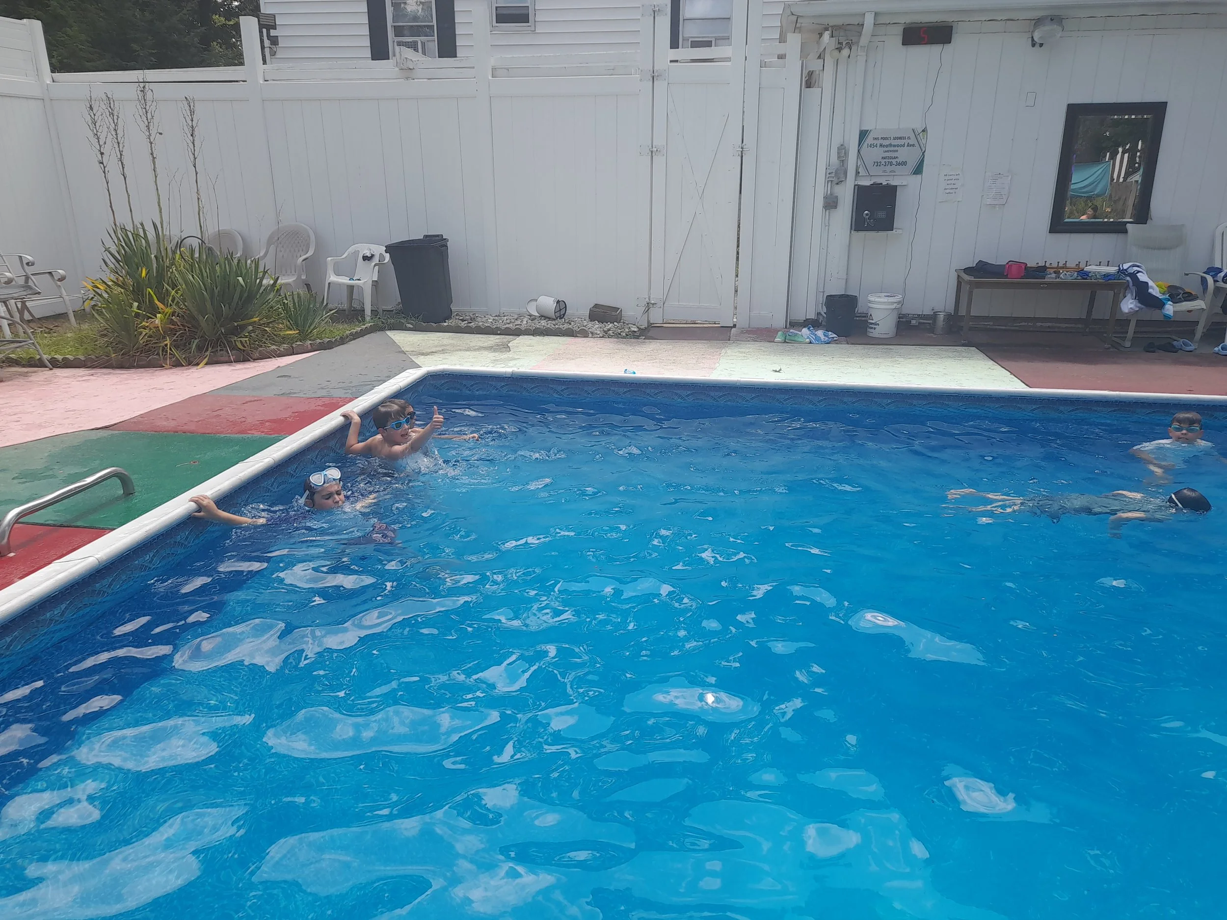 Children swimming and playing in a residential outdoor pool with a white fence and poolside seating in the background.