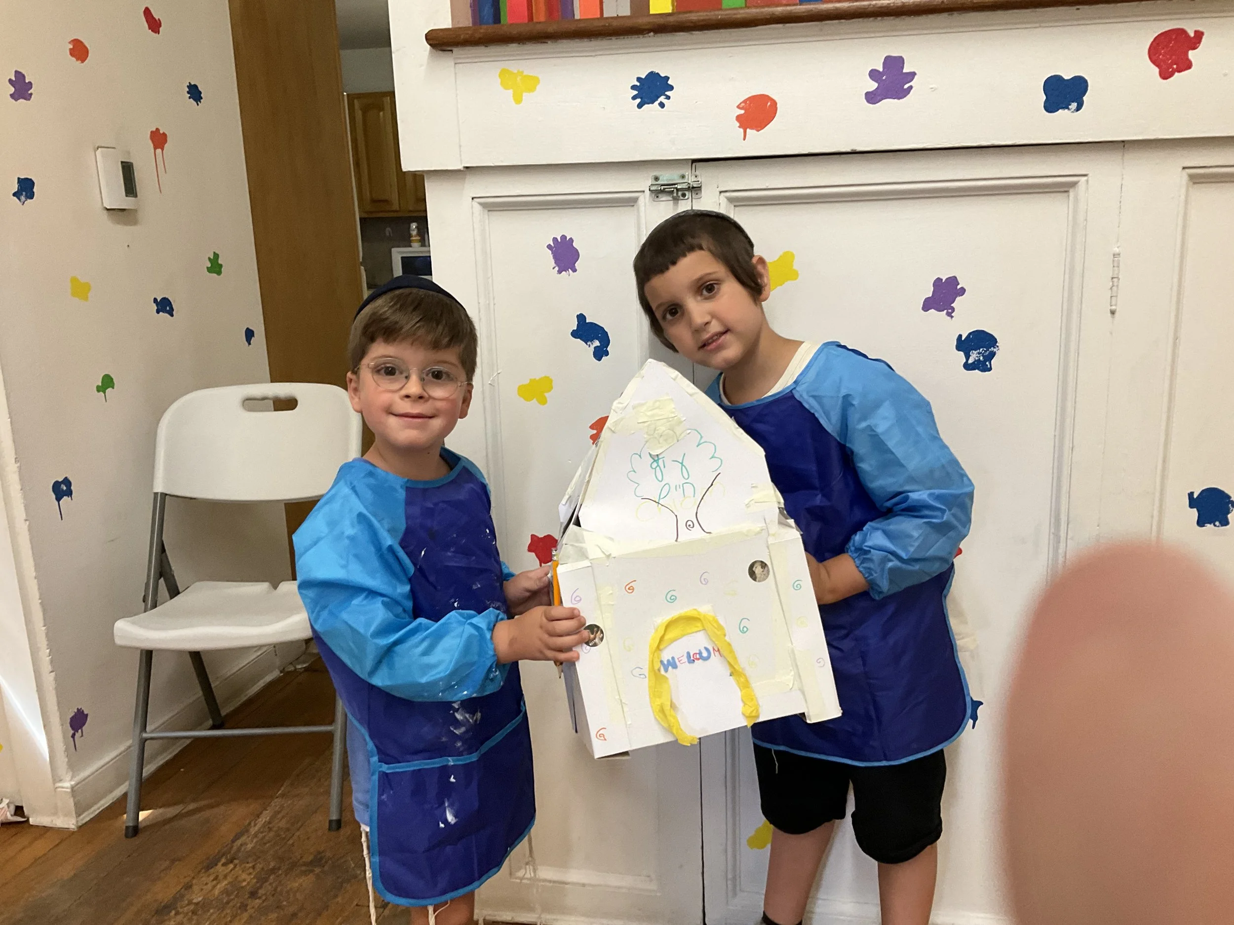 Two children wearing blue art smocks holding a homemade cardboard project with drawings, standing next to a white wall decorated with colorful paint splatters.