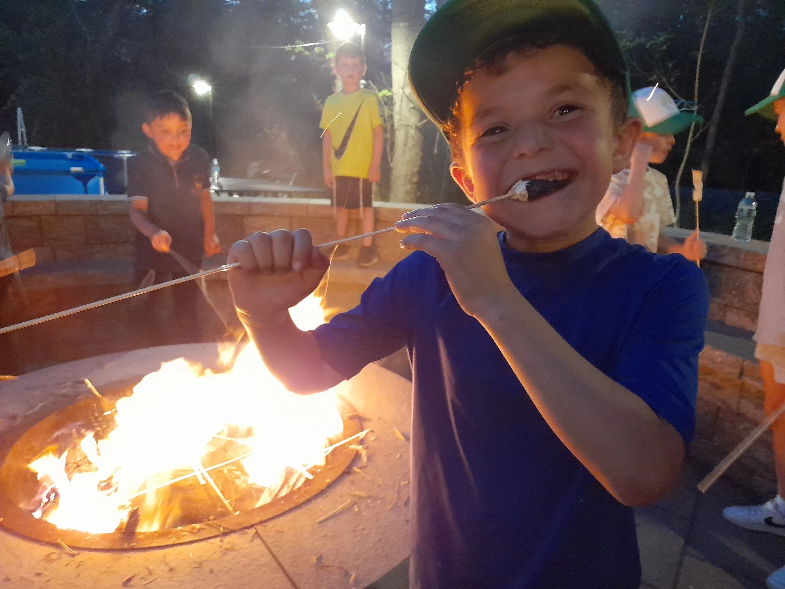 Children roasting marshmallows over a fire pit in the evening, with one child smiling in the foreground holding a roasted marshmallow on a stick.