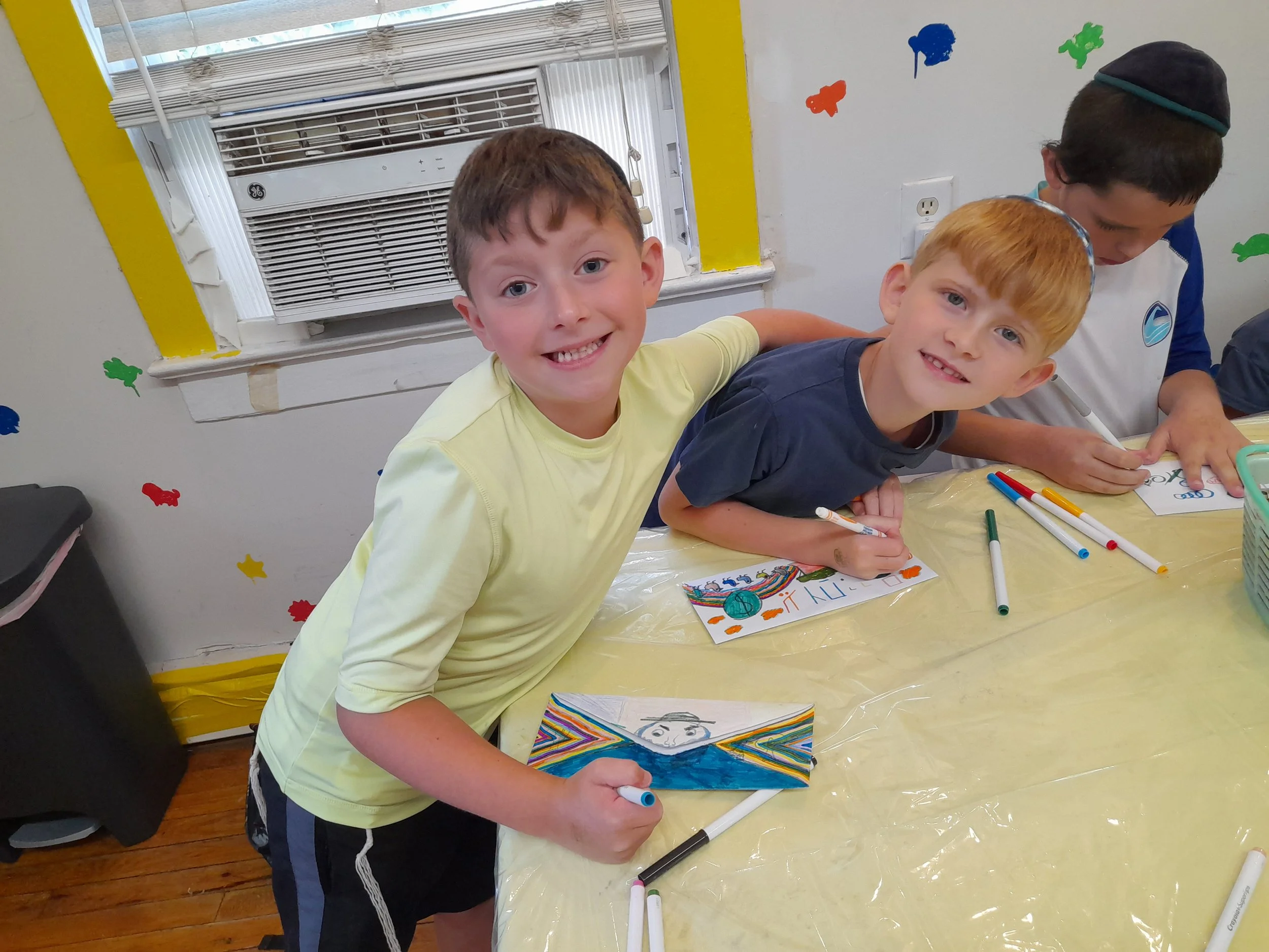 Children engaged in arts and crafts, drawing on paper with markers, in a brightly decorated room with colorful wall stickers and an air conditioner in the background.