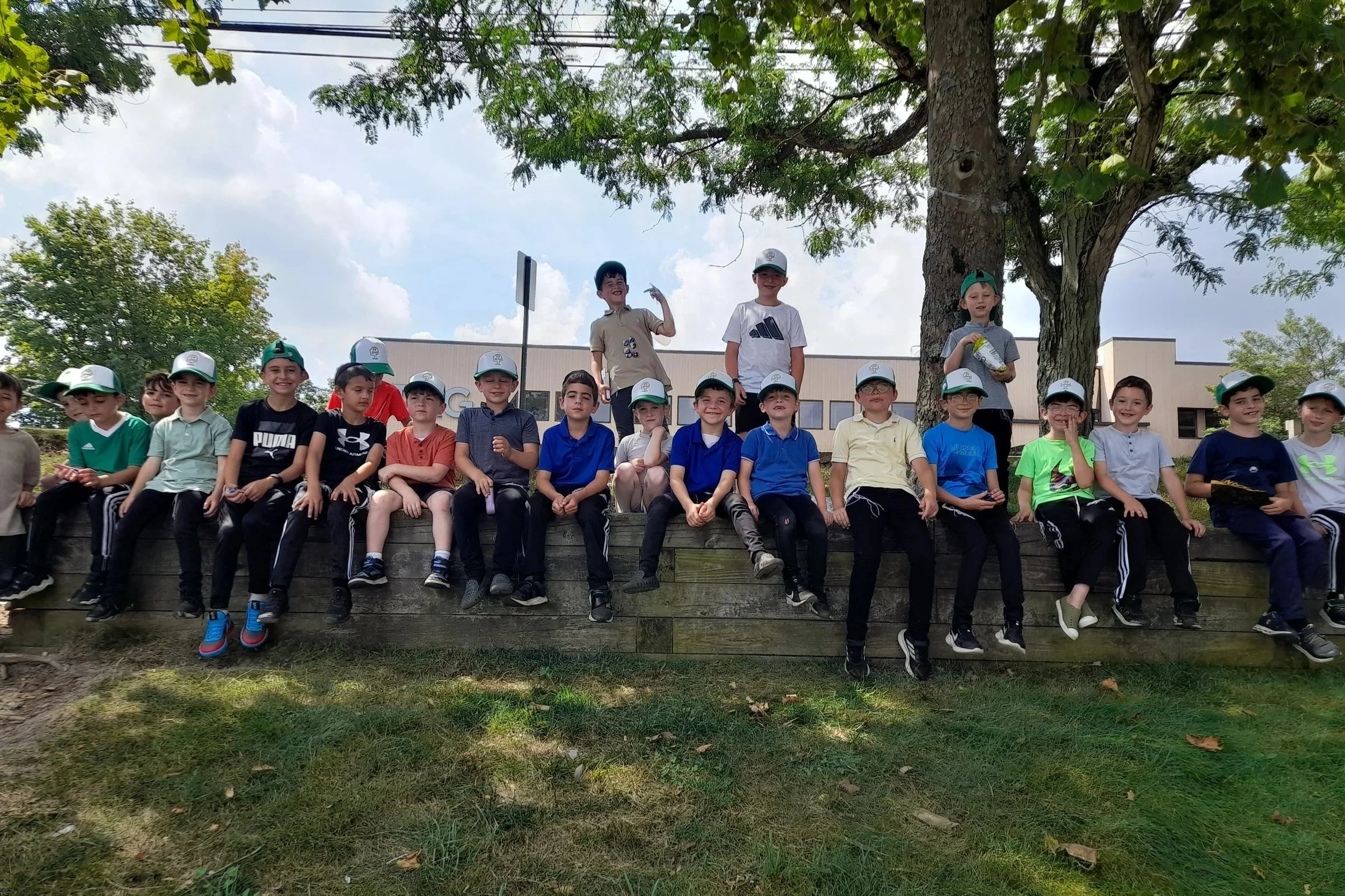 Group of children sitting on a wooden ledge outdoors under a large tree, some wearing hats, with a building in the background on a partly cloudy day.