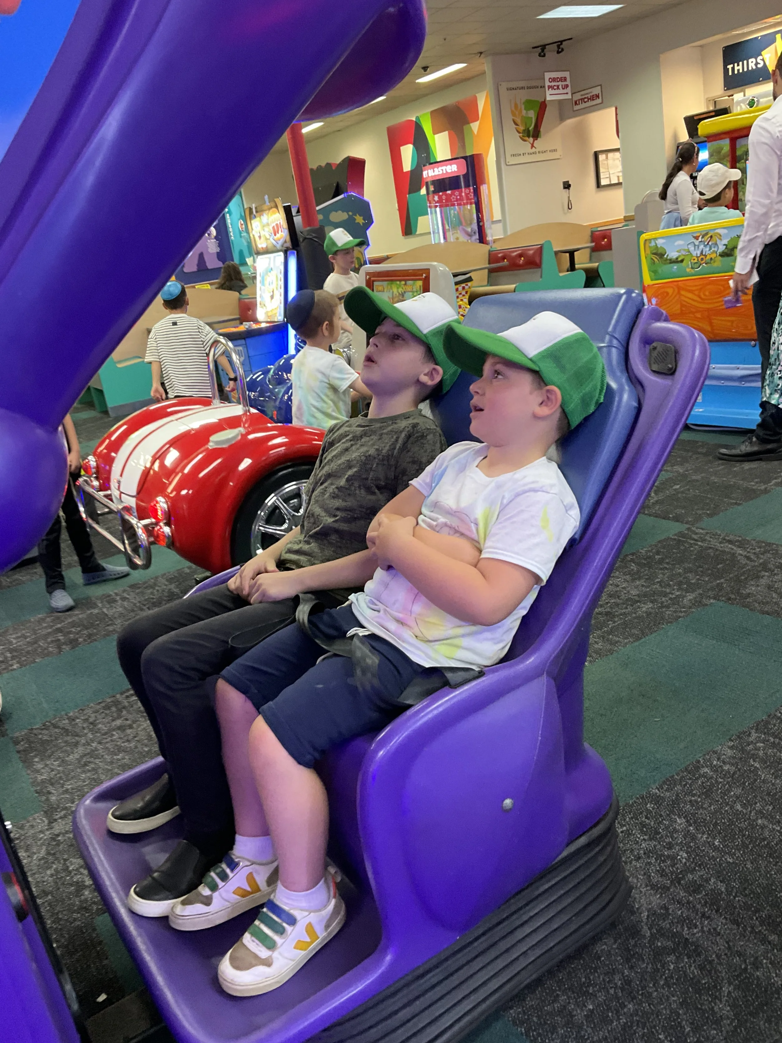 Children sitting in an arcade ride, wearing green and white caps, surrounded by colorful gaming machines and other children.