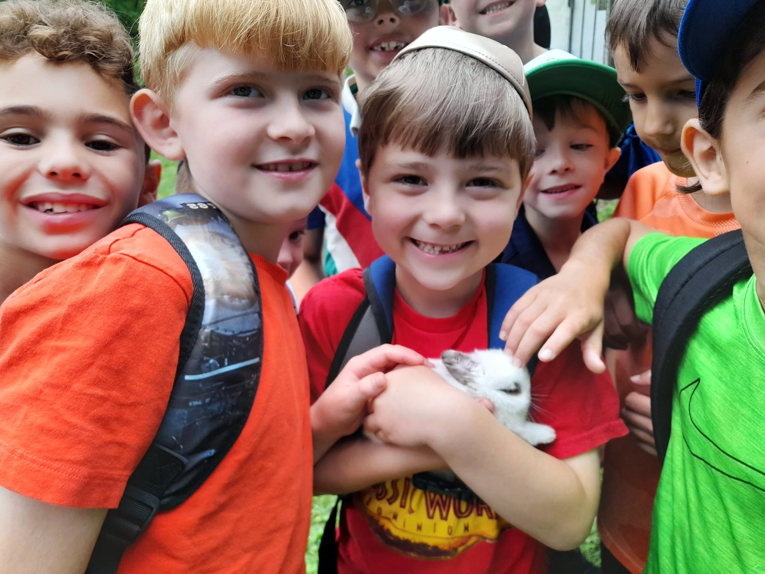 A group of smiling children, mostly boys, surround and hold a small white rabbit outdoors. They are wearing colorful shirts and backpacks, suggesting a school or camp setting.