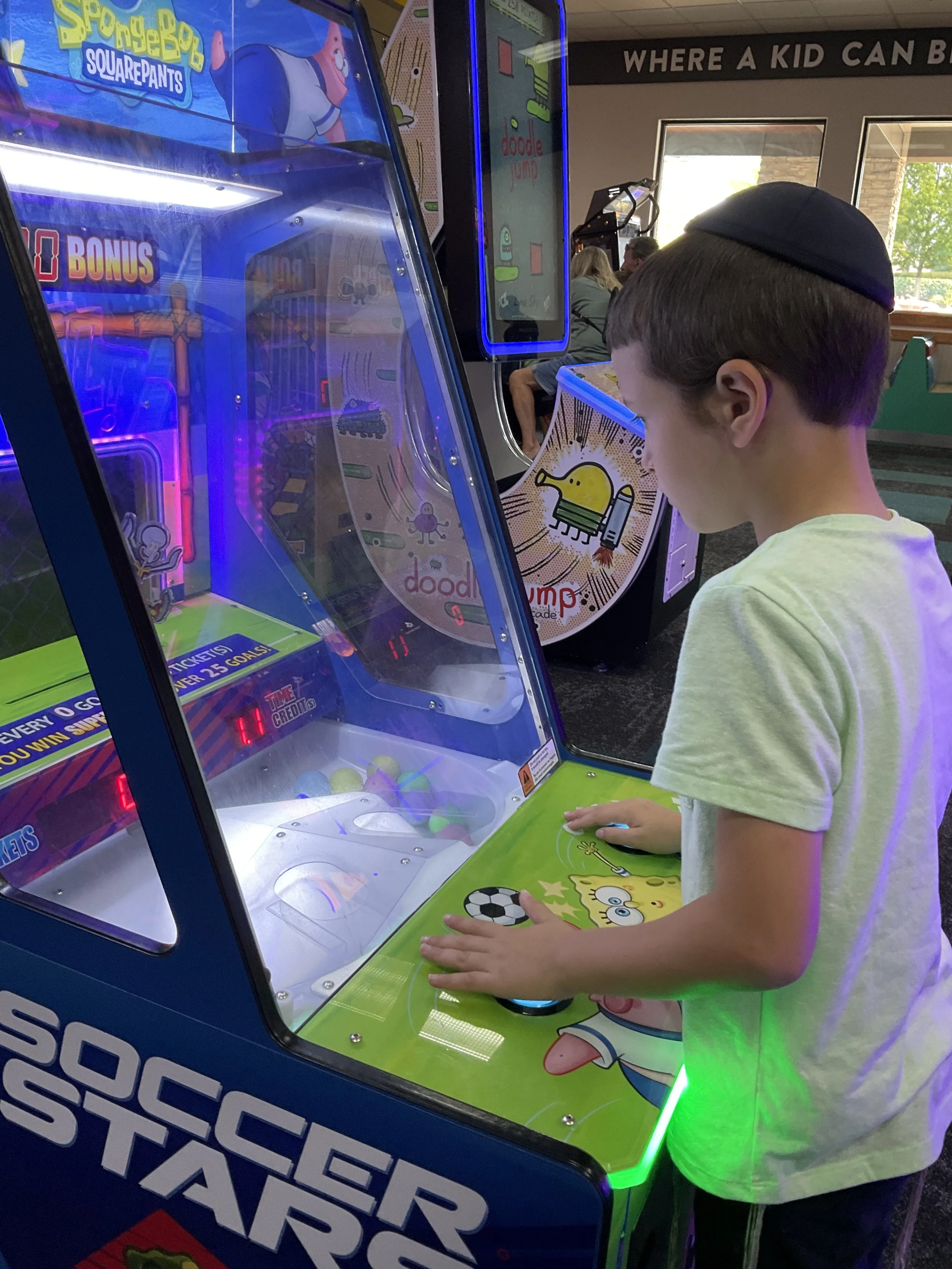 Child playing an arcade game in a game center, featuring Spongebob Squarepants graphics. Another game machine, Doodle Jump, is visible in the background.