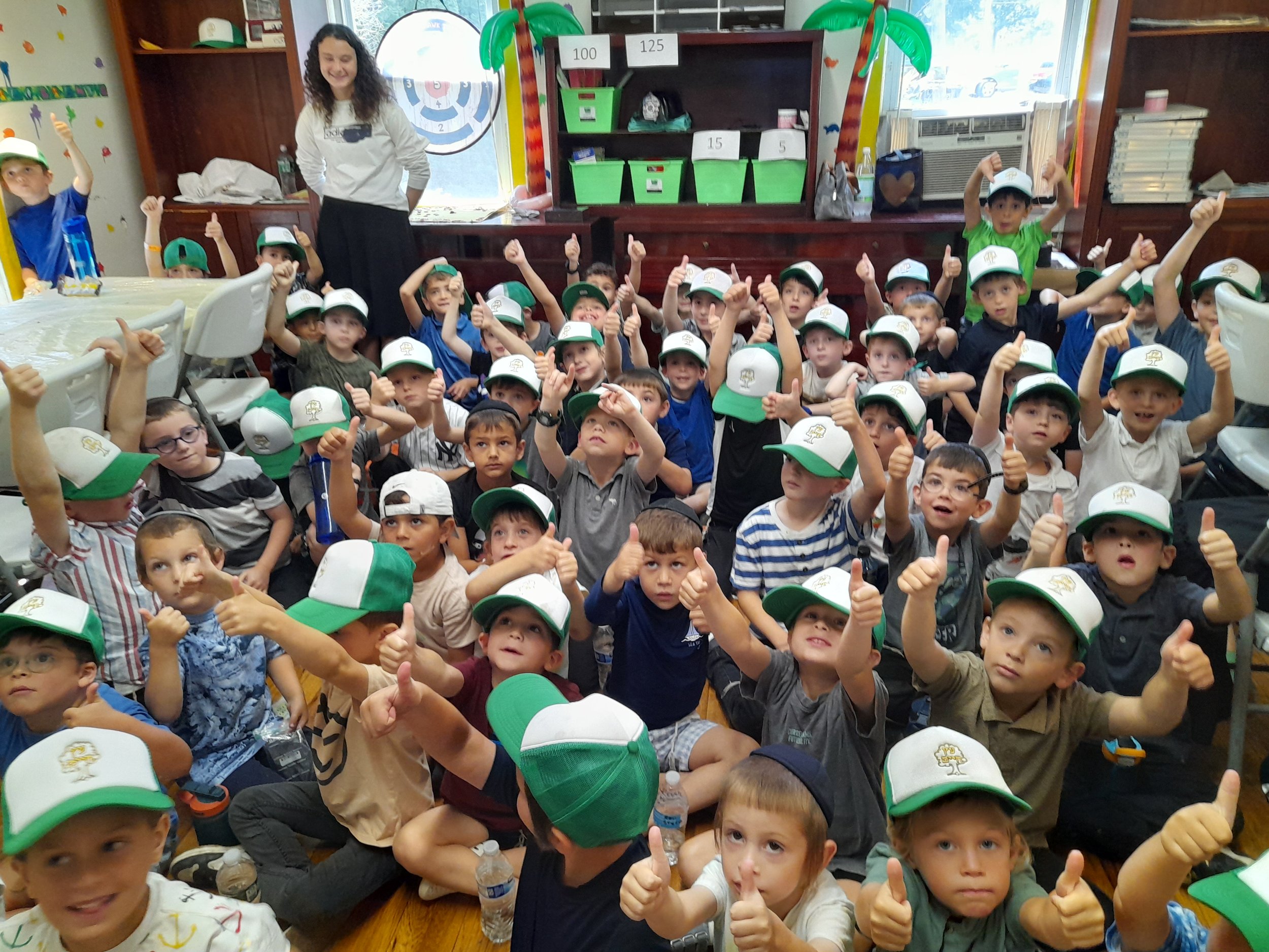 Group of children wearing green and white hats giving thumbs up in a classroom with a teacher standing behind them.