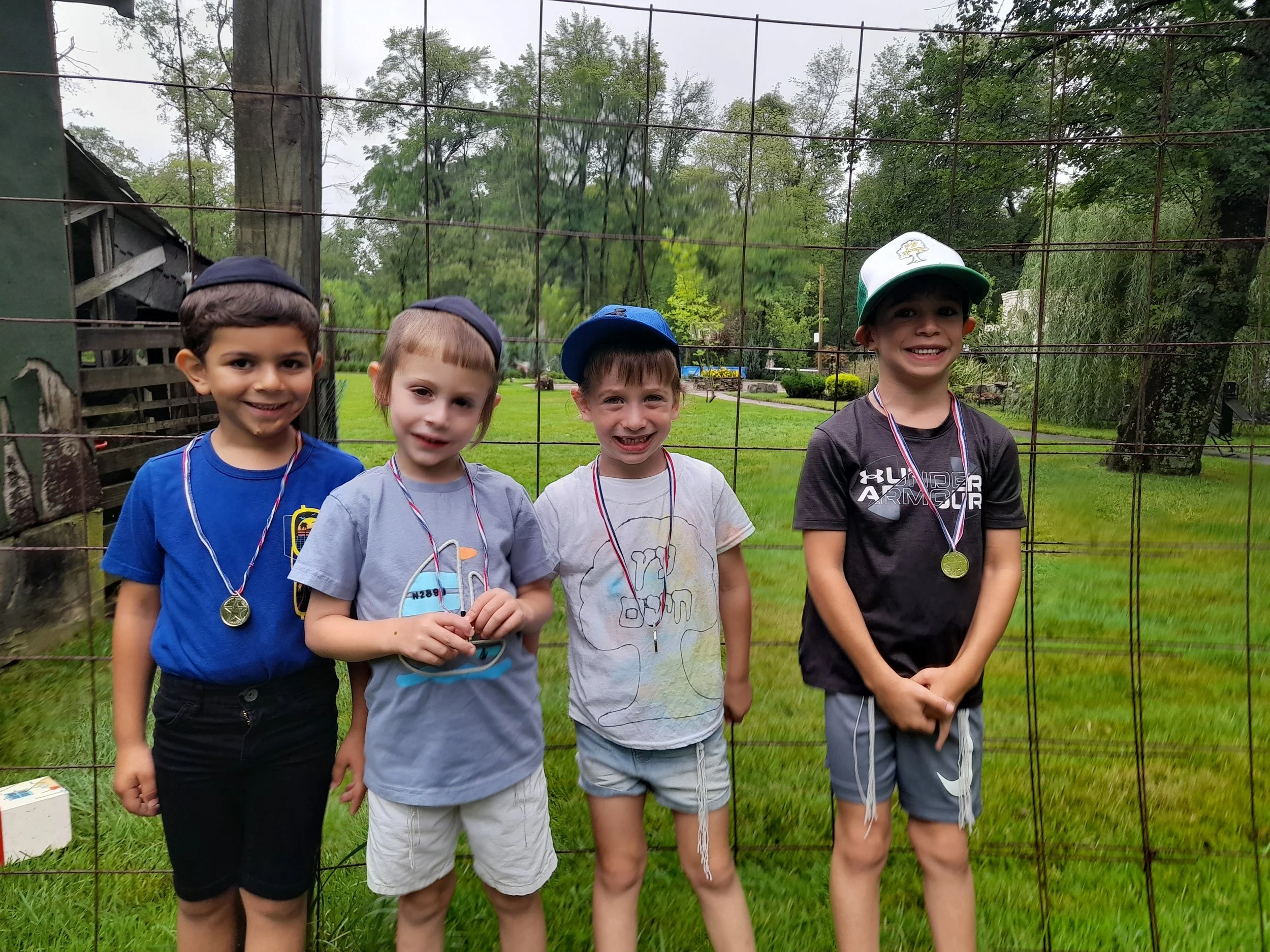 Four young boys smiling, wearing medals and caps, standing outdoors in front of a wire fence, with grassy area and trees in the background.