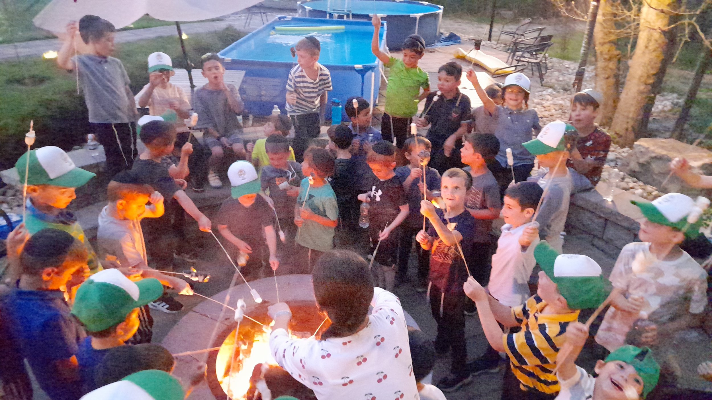 A group of children roasting marshmallows over a campfire, with some children wearing green and white caps. Behind them, there's a blue pool and a seating area. It's an outdoor gathering likely in the evening.