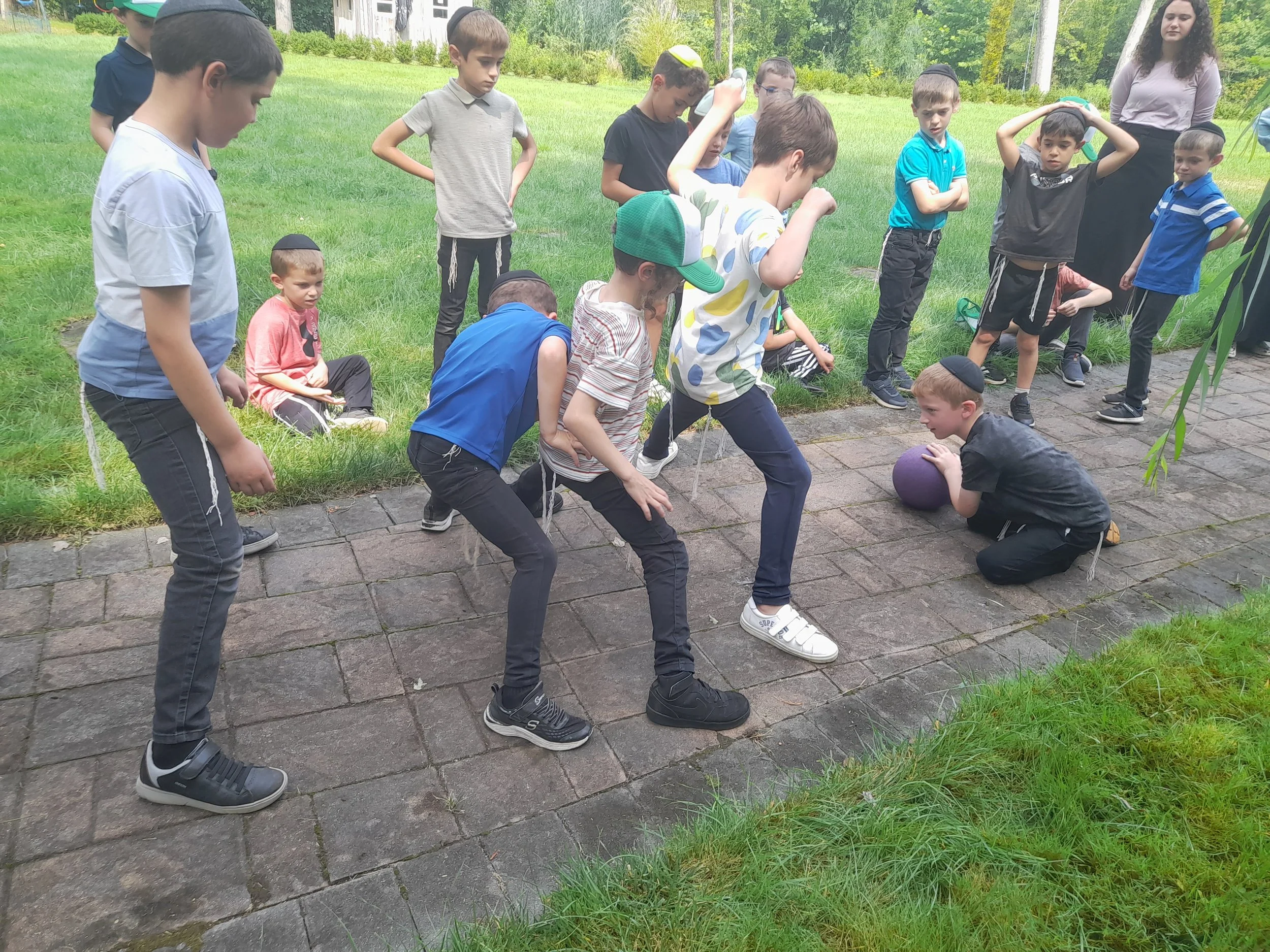 Group of boys playing a game outdoors on a paved walkway, surrounded by grass. One boy is kneeling with a ball while others are observing closely. Trees and a building are in the background.