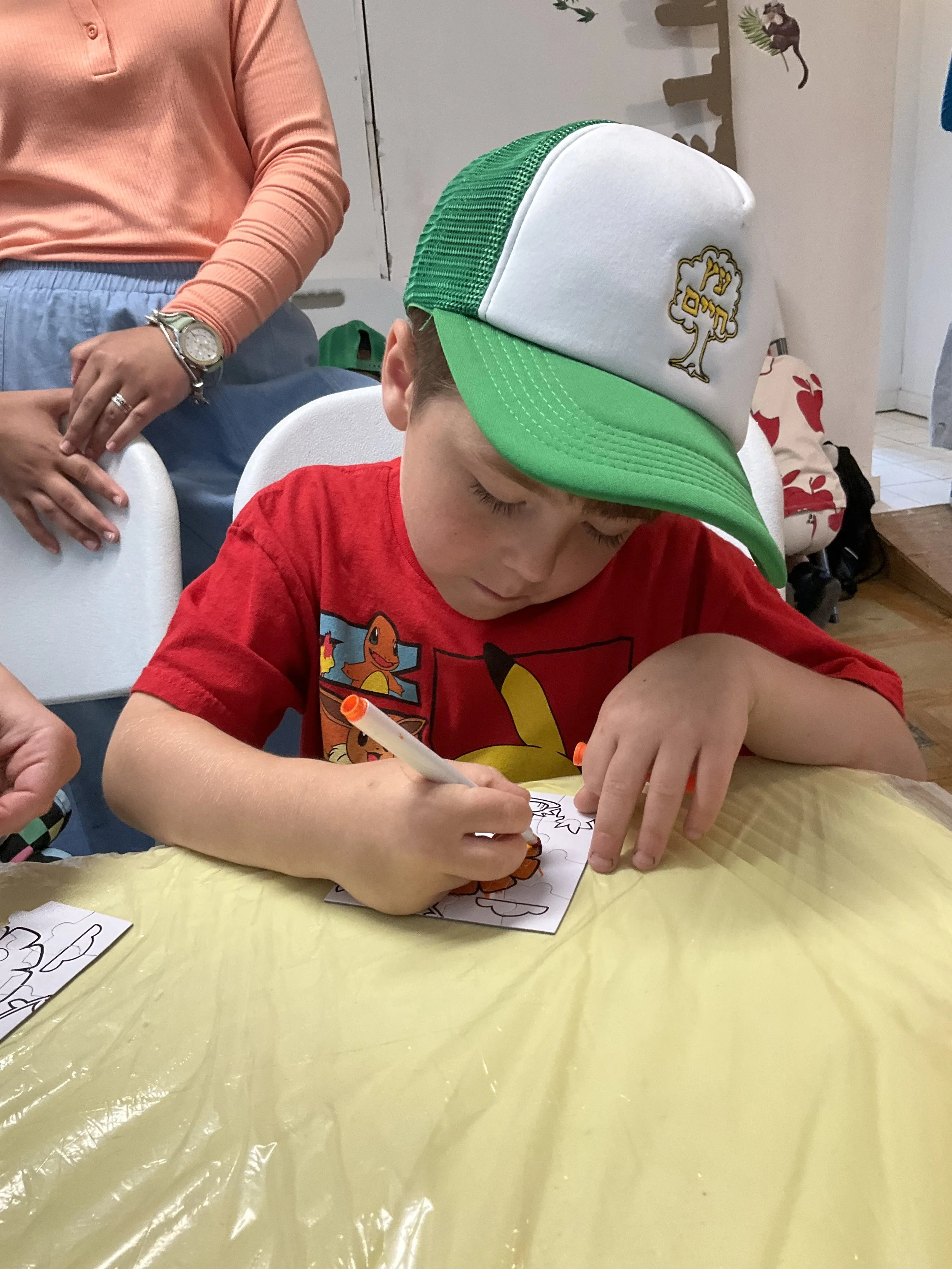 Young boy coloring with a marker while wearing a green and white cap and a red shirt featuring a cartoon design.