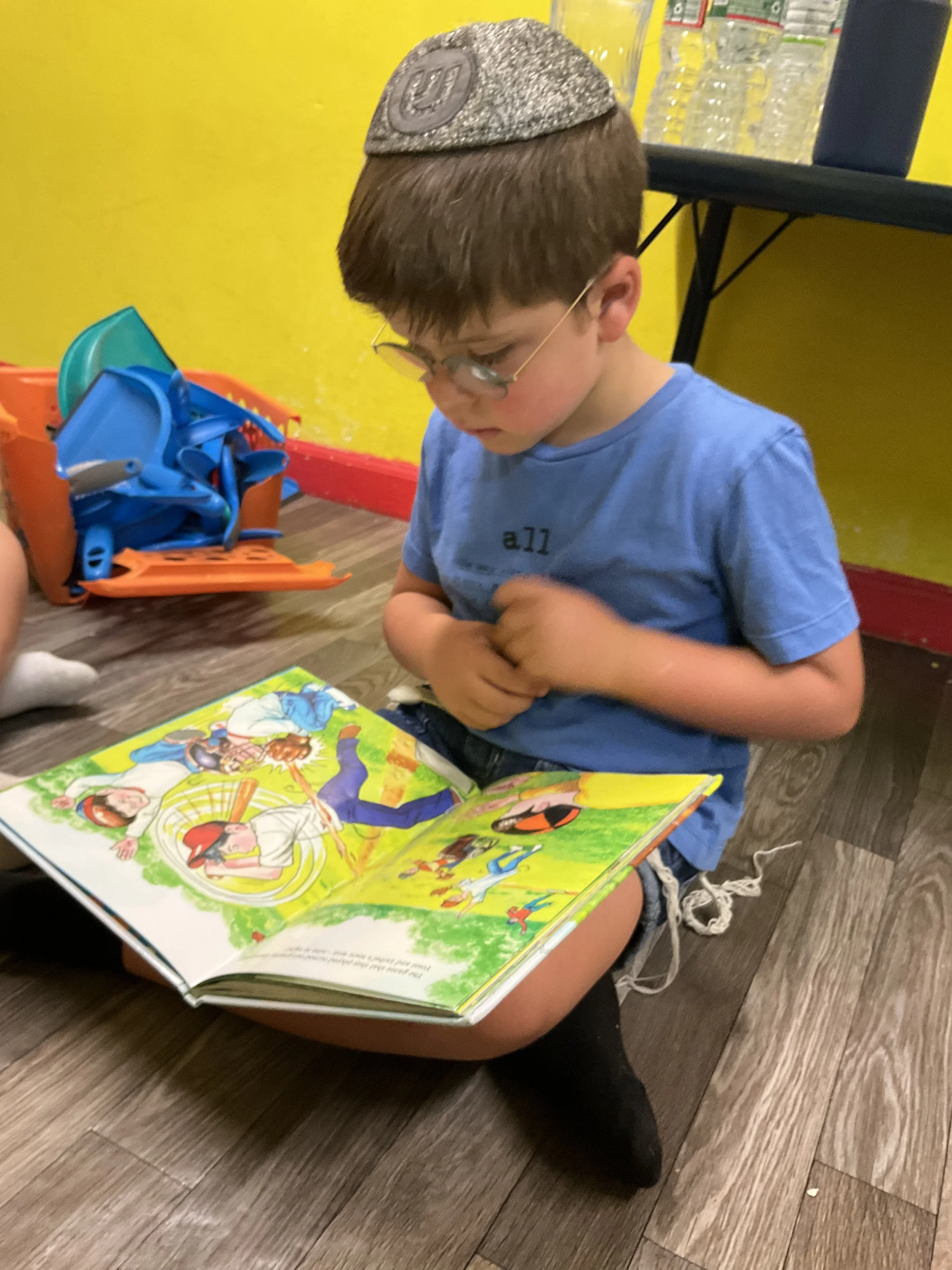Child reading a colorful storybook while sitting on the floor, wearing glasses and a kippah. In the background, a yellow wall and blue and orange stackable seats are visible.
