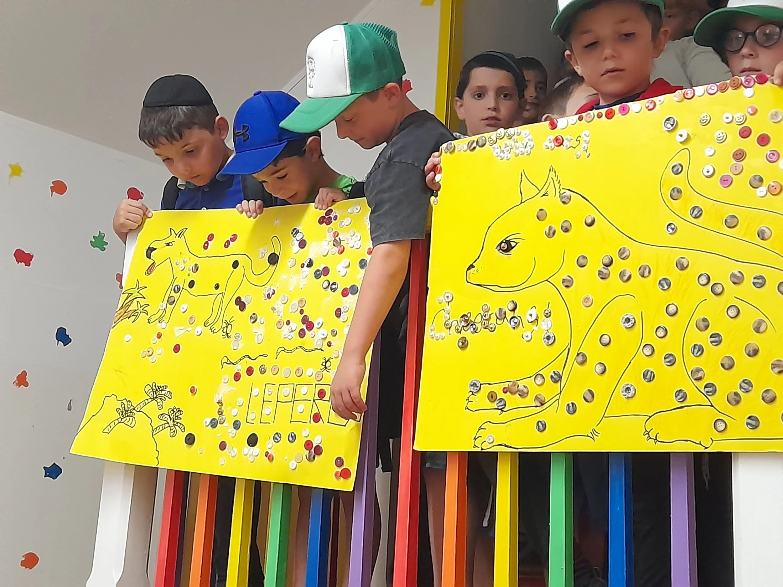 Children holding colorful posters decorated with button art featuring animals, possibly a dog and a large cat, on a yellow background.