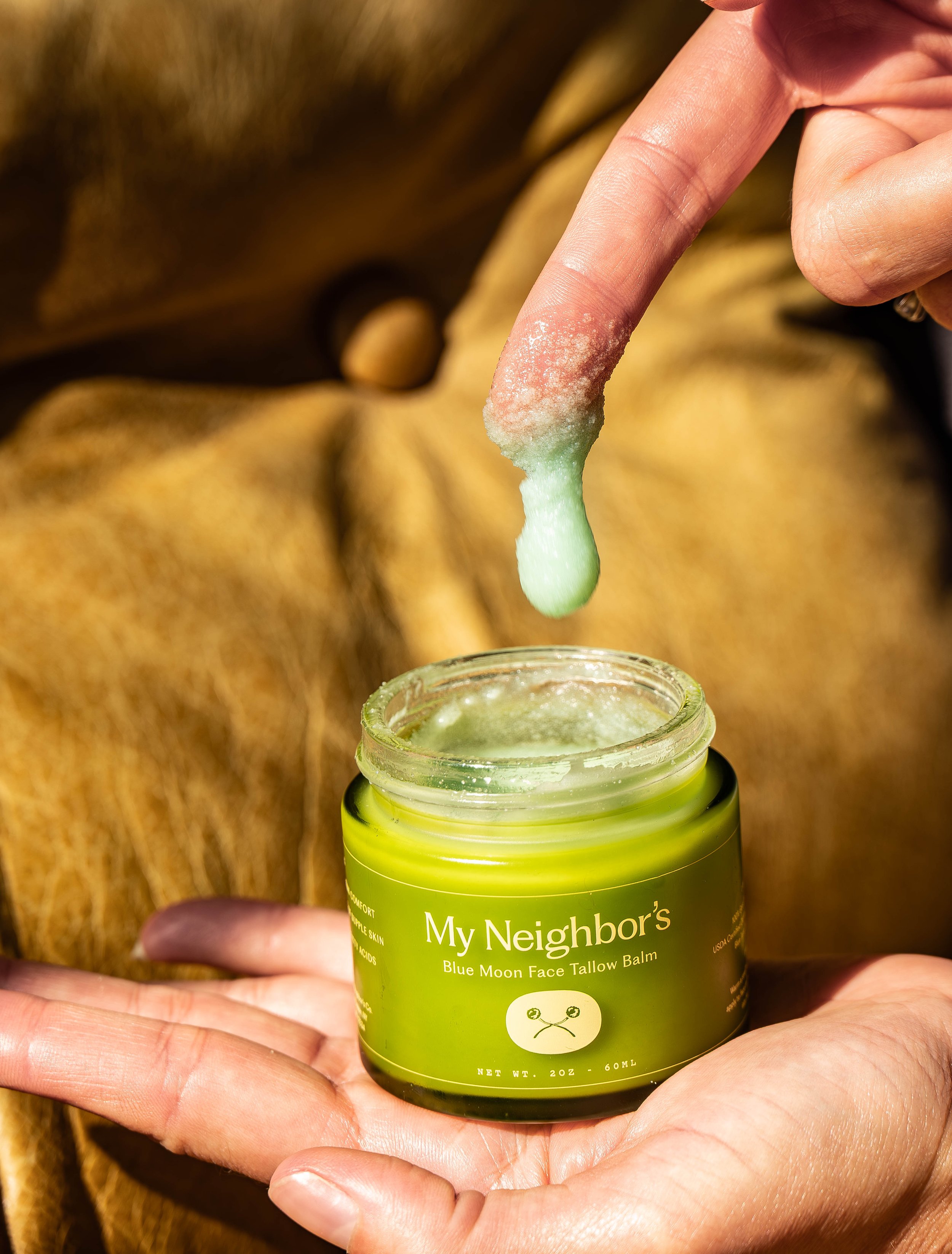 Close-up of a hand holding a green jar labeled "My Neighbor’s Balm, Blue Moon Face Tallow Balm" with a finger dipped into the balm, which is a light green, creamy substance.