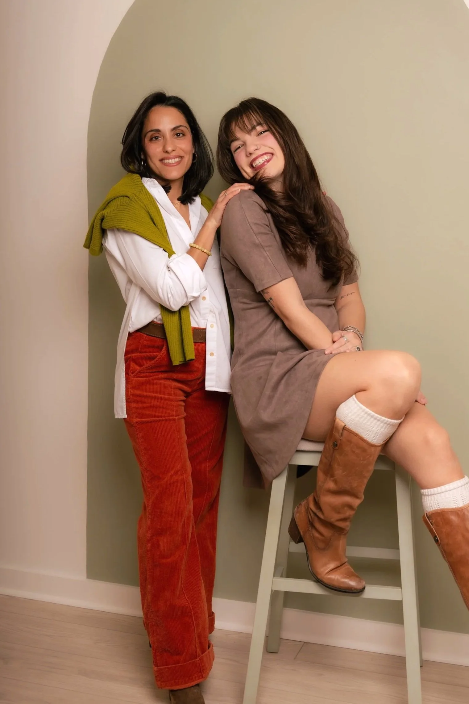 Abby Fritsch, brand designer and Ariel Lomeli, founder, standing and one sitting on a stool, in a room with light-colored walls.