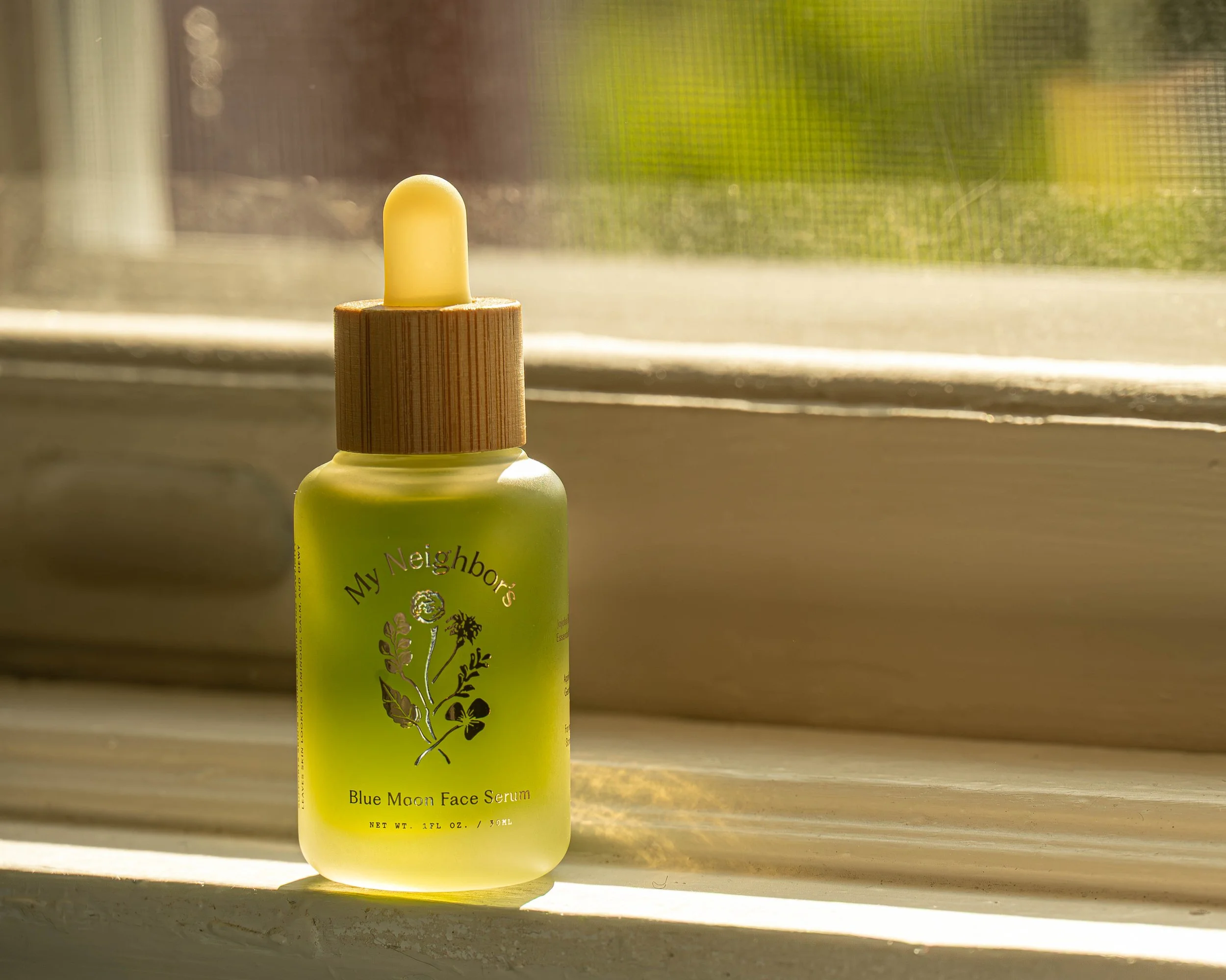 A small bottle of My Neighbor's Blue Moon Face Serum with a wooden cap, placed on a windowsill with sunlight streaming in and a blurred green outdoor background.