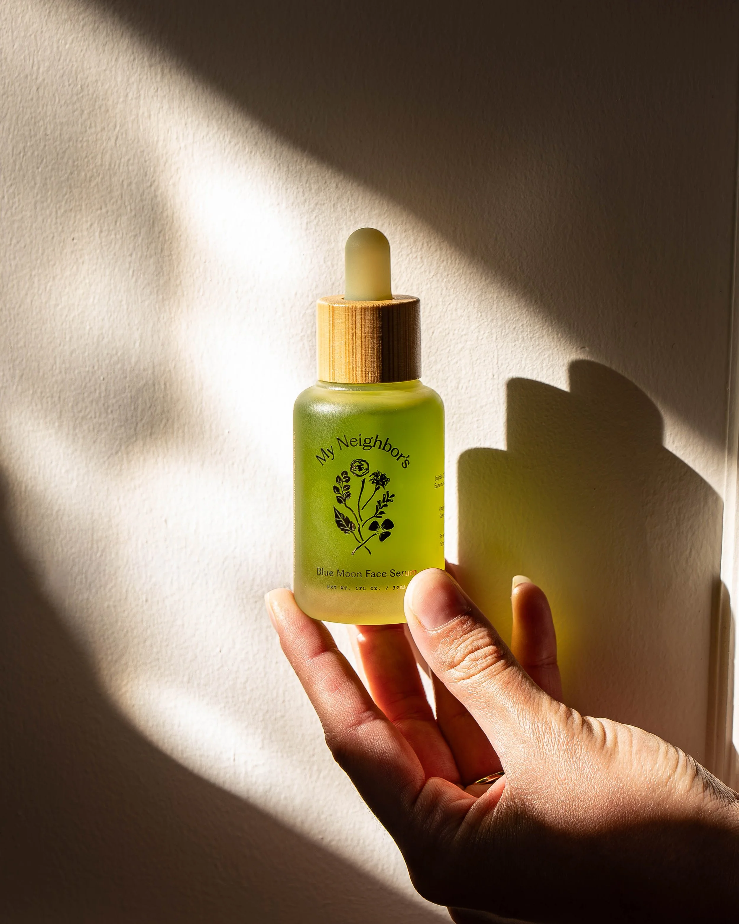 Close-up of a hand holding a green bottle of face serum with a wooden dropper cap, against a background wall with lighting creating shadow effects.