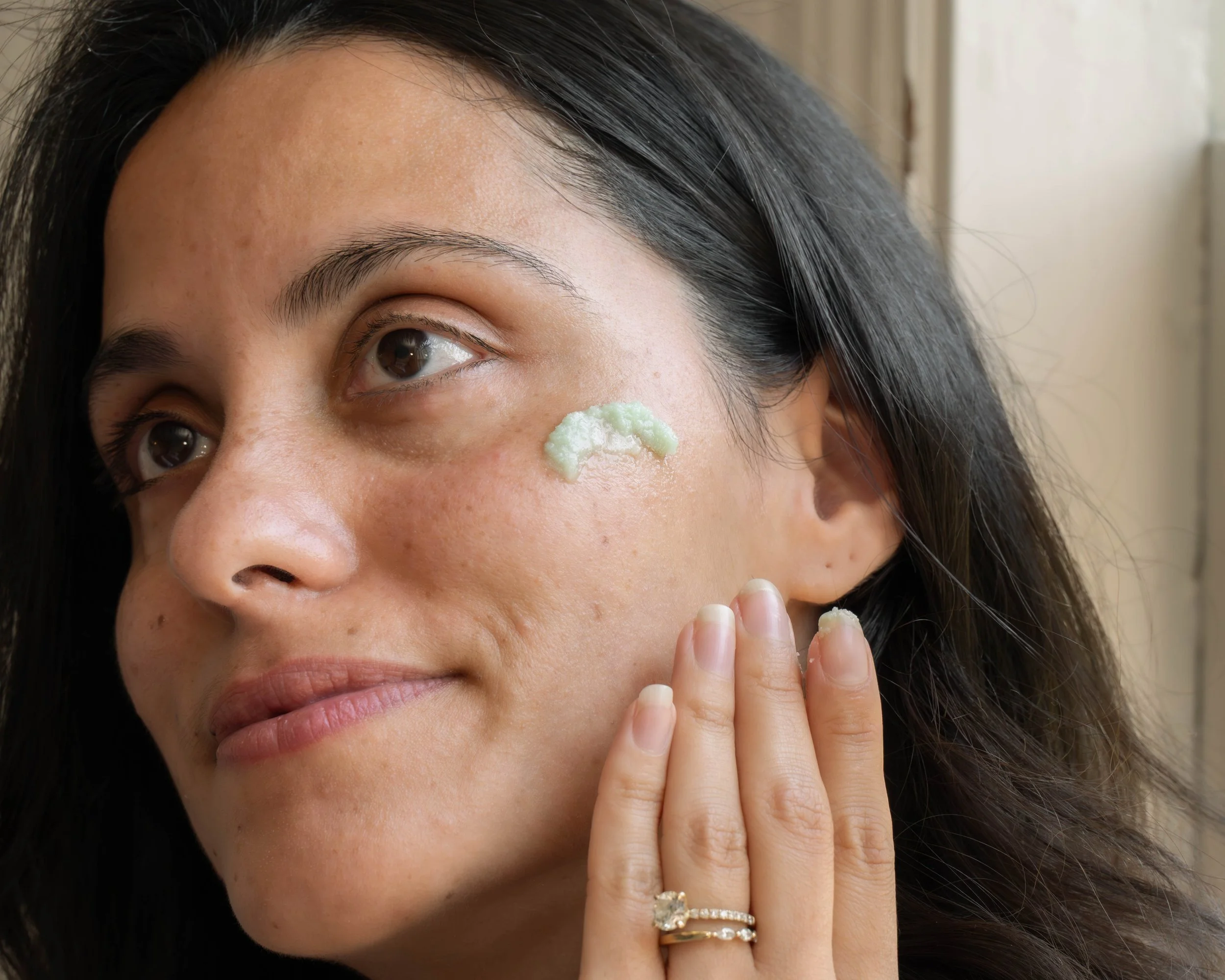 A woman with long dark hair applying a green facial mask to her cheek with her fingers.