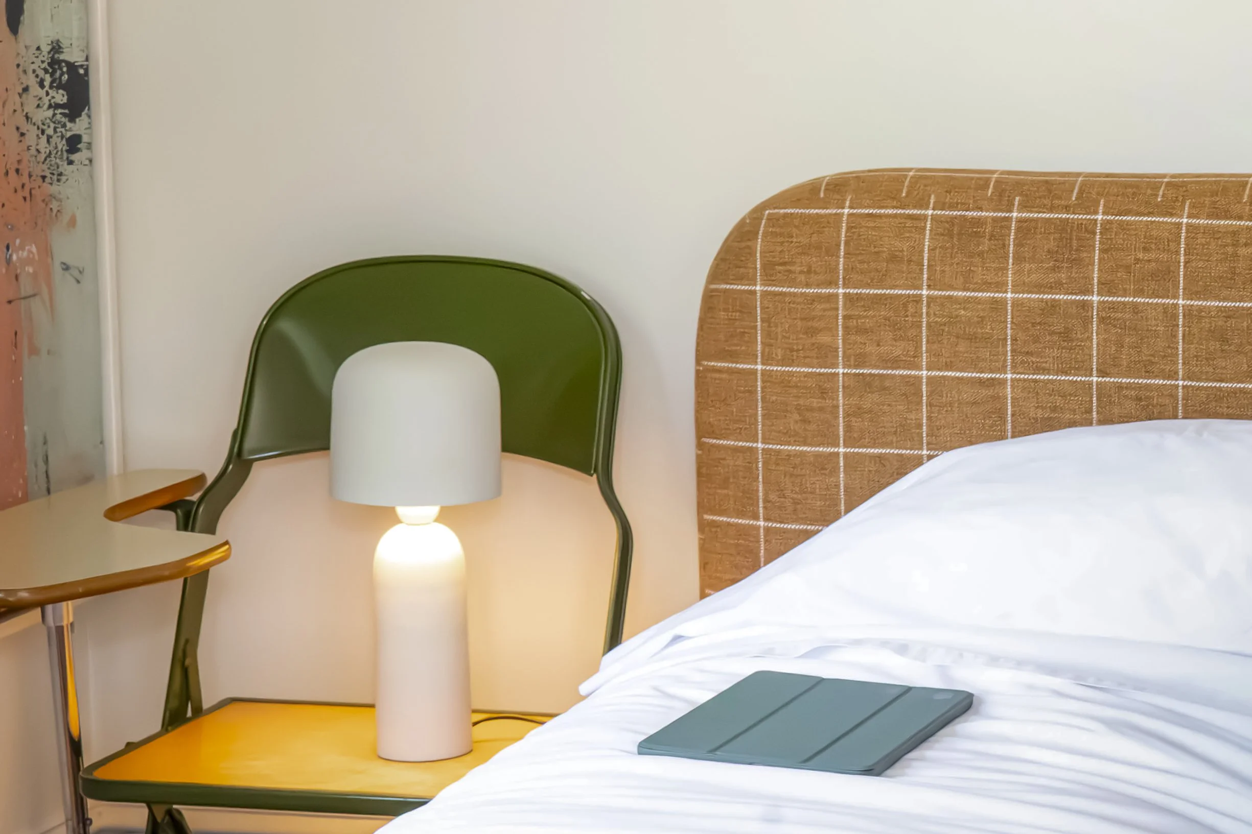 Interior of a room with a bed, a wooden bedside table, a green chair, a white lamp, and a notebook on the bed. The wall behind has a headboard with a brown and white checked pattern.