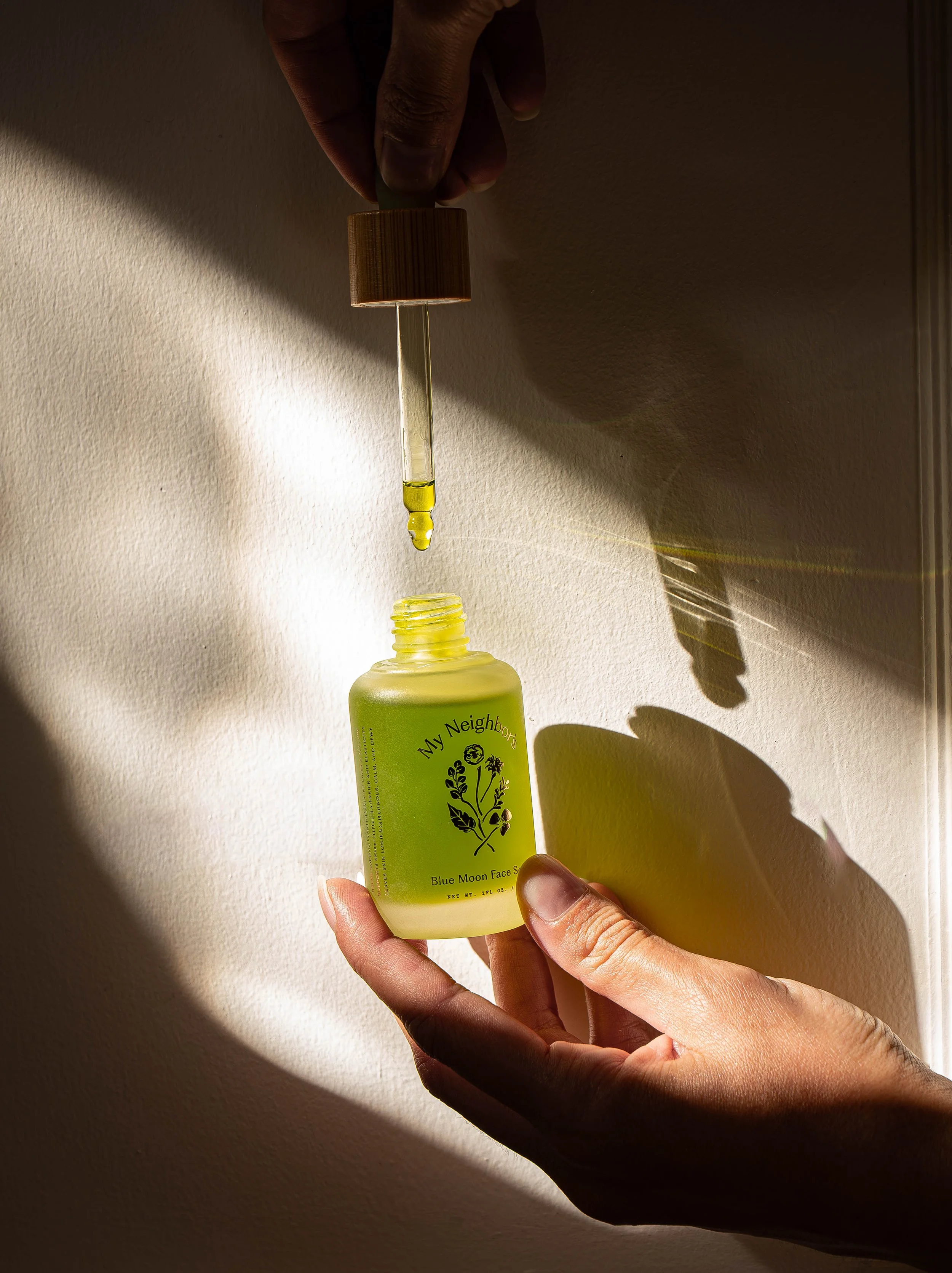 A person holds a yellow bottle labeled 'My Neighbors' and is using a dropper to transfer a yellow liquid onto a yellow dish, with shadows cast on a textured wall.