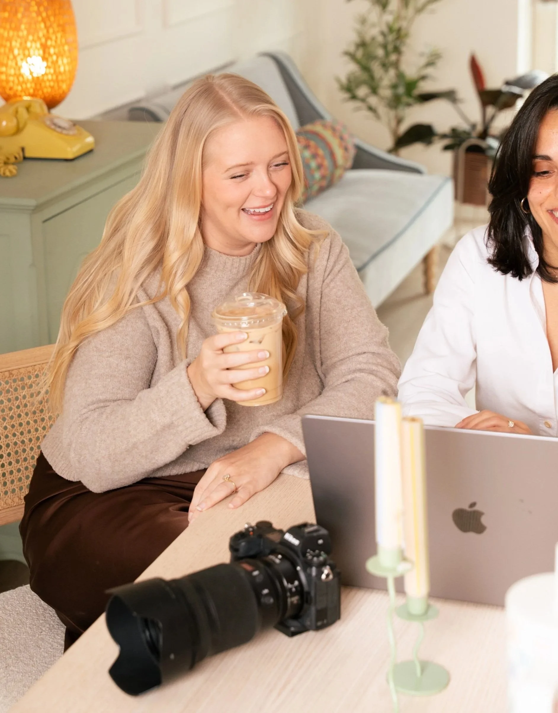 Caitlin Holbrook, photographer at Camille Maede, holding an iced coffee, sitting at a table in a cozy home, with a camera and a laptop on the table, and smiling.