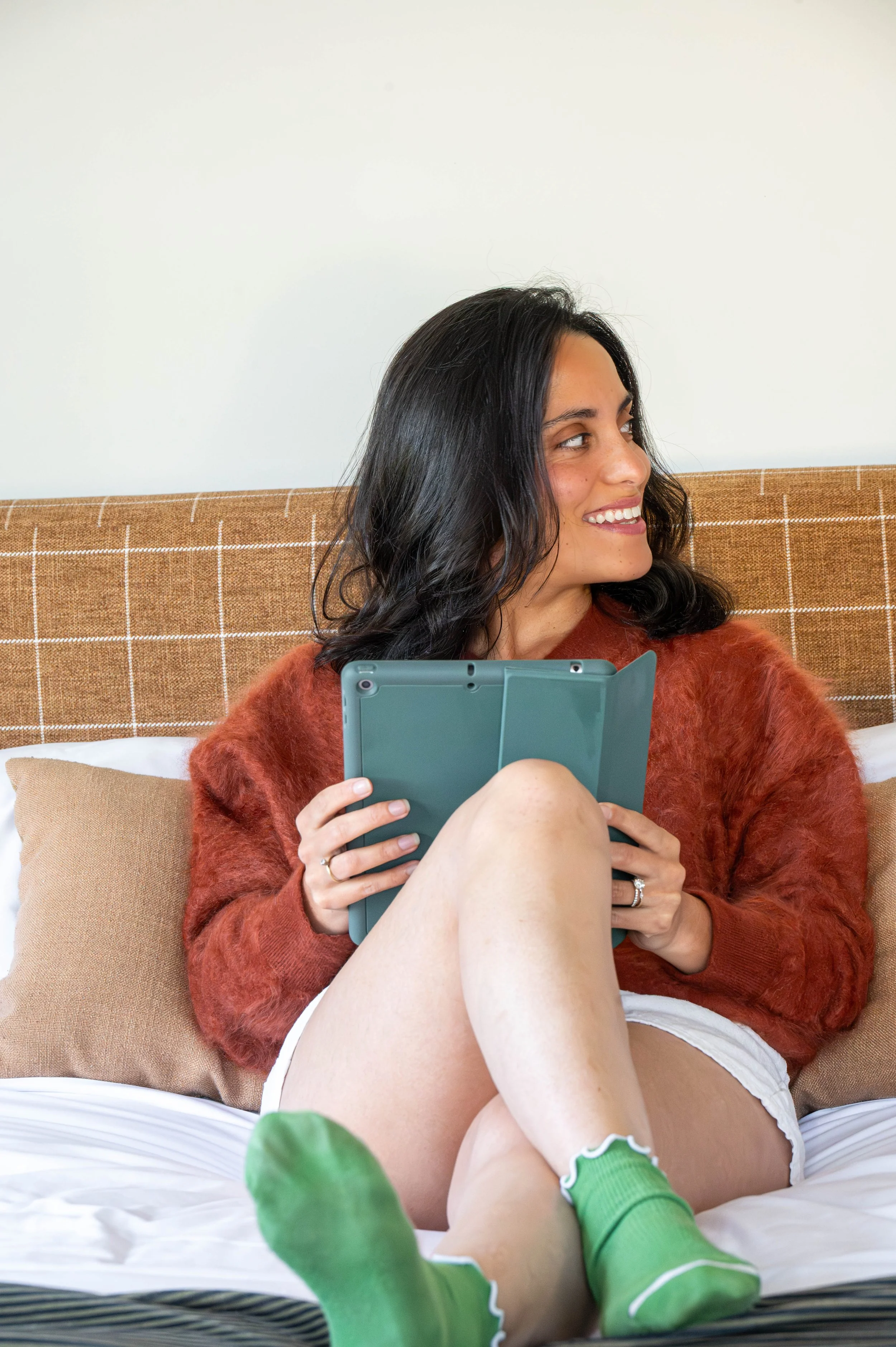 Ariel is holding a tablet. She is wearing a rust-colored fuzzy sweater and white shorts with bright green socks. The bed has a checkered headboard and beige pillows, with a colorful blanket partially visible.