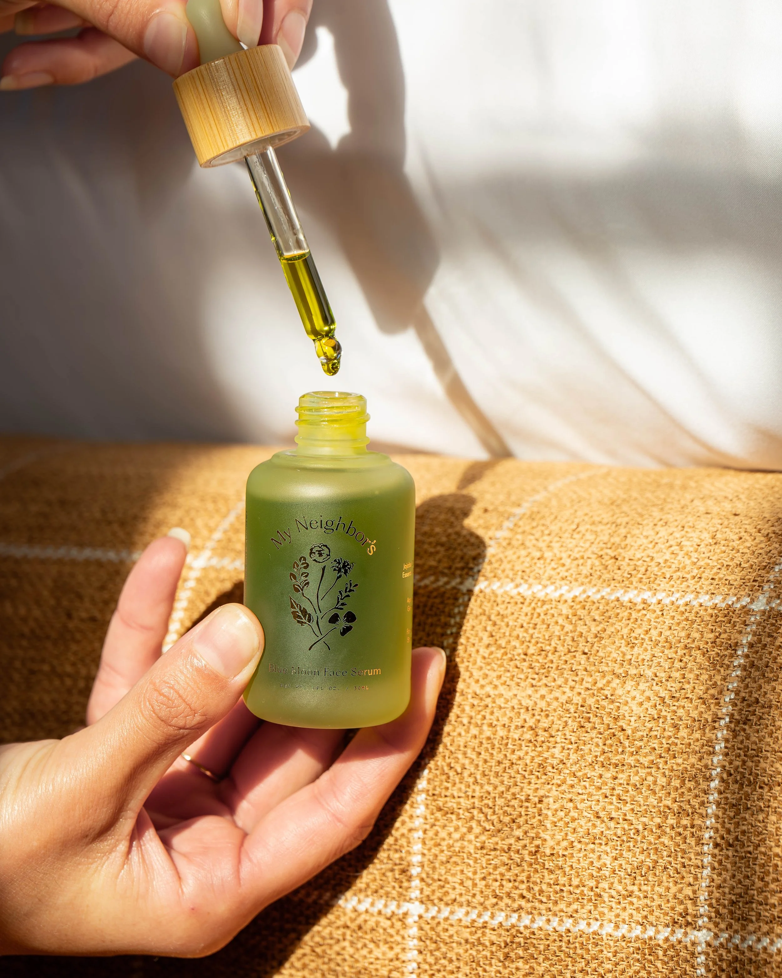 A person's hand holding a dropper with yellow serum above a small green bottle on a textured beige surface. The serum is being dispensed from the dropper into the bottle opening.