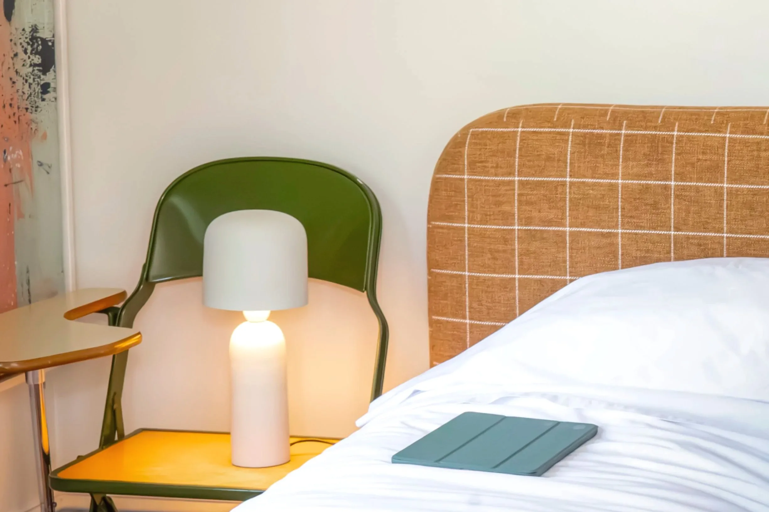 A bedroom scene with a portion of a bed with white bedding and a tablet on it. Next to the bed, there is a yellow bedside table with a modern lamp on top. Two chairs are visible, one green and one wooden, positioned near the table. A lamp on the table illuminates the area. The background has a white wall and a partial view of a colorful abstract artwork on the wall.