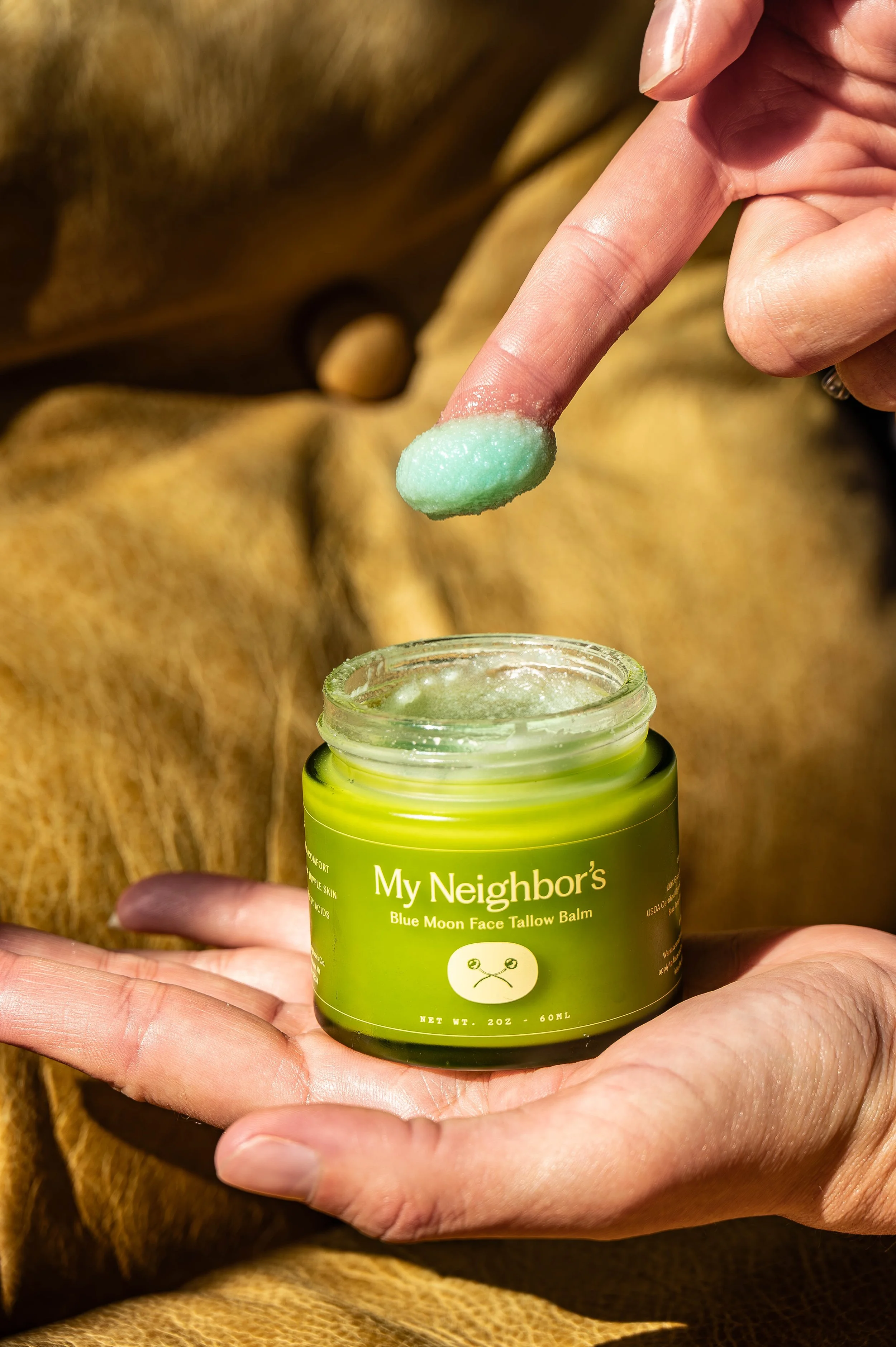 A person holding a small green jar labeled 'My Neighbor's Blue Moon Face Tallow Balm' with a finger dipping a green balm from the jar.